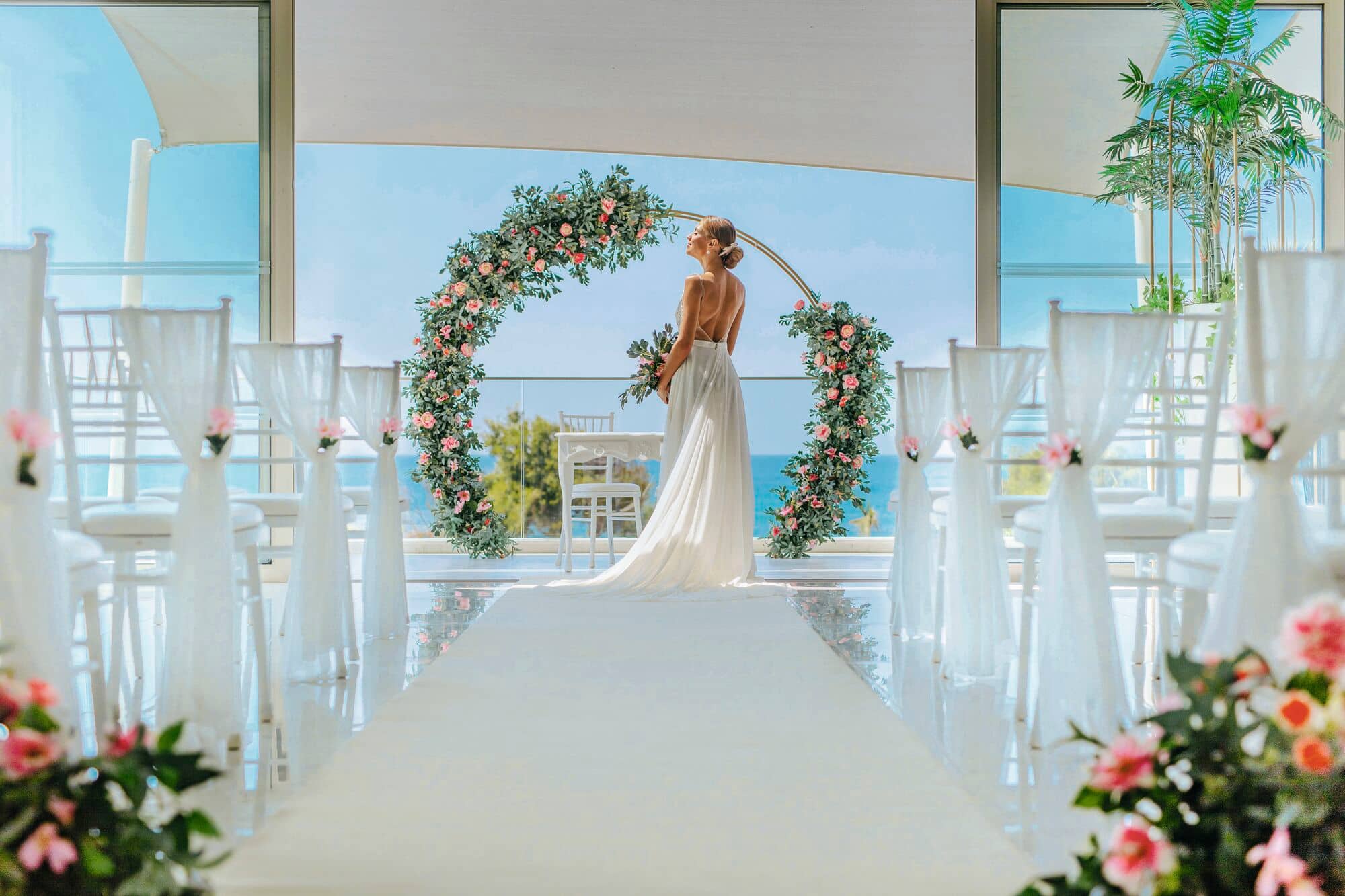 An impressive panoramic view out to sea lies behind the ceremony. Flowers and white chairs line the aisle, looking toward a bride and a striking floral arrangement.