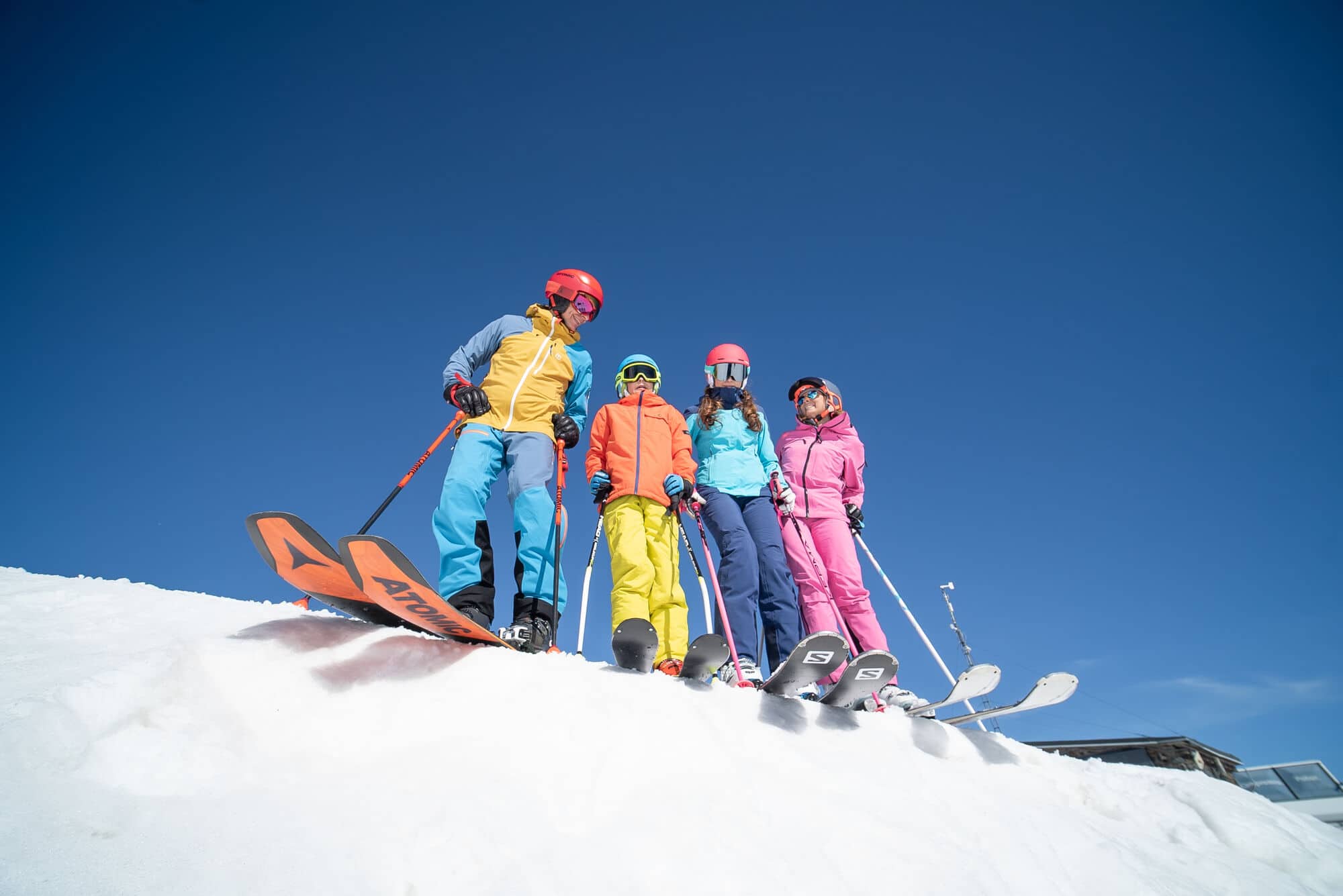 Family of skiers in colourful winter gear standing on a snowy slope under a clear blue sky.