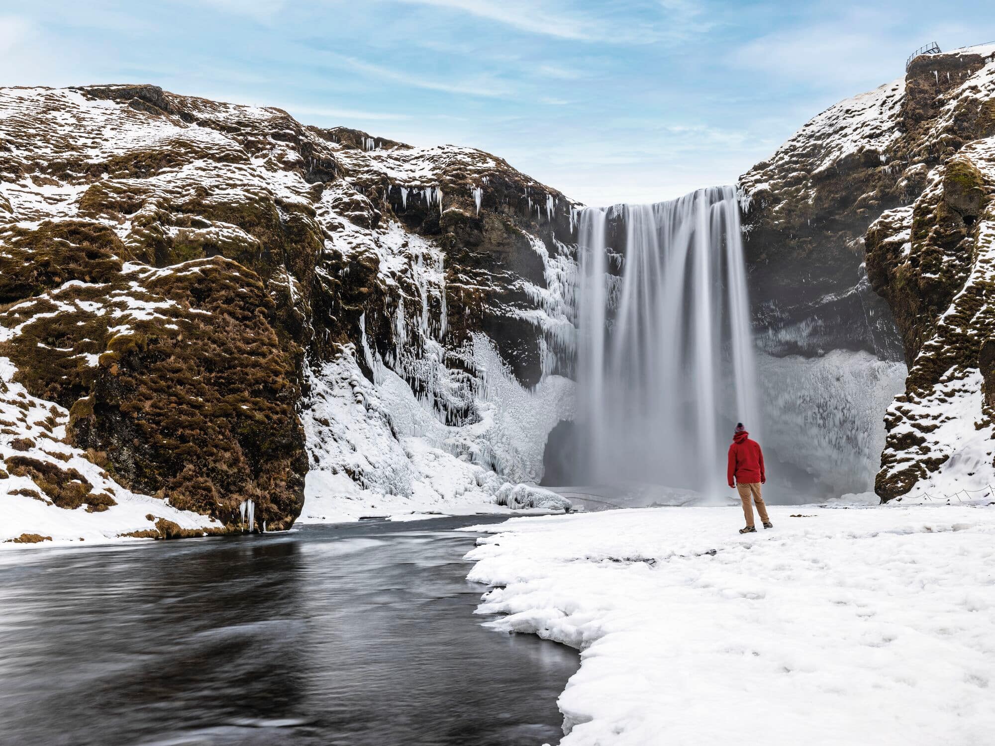 A person wearing a red jacket stands on snow-covered ground near a partially frozen waterfall surrounded by rocky cliffs.