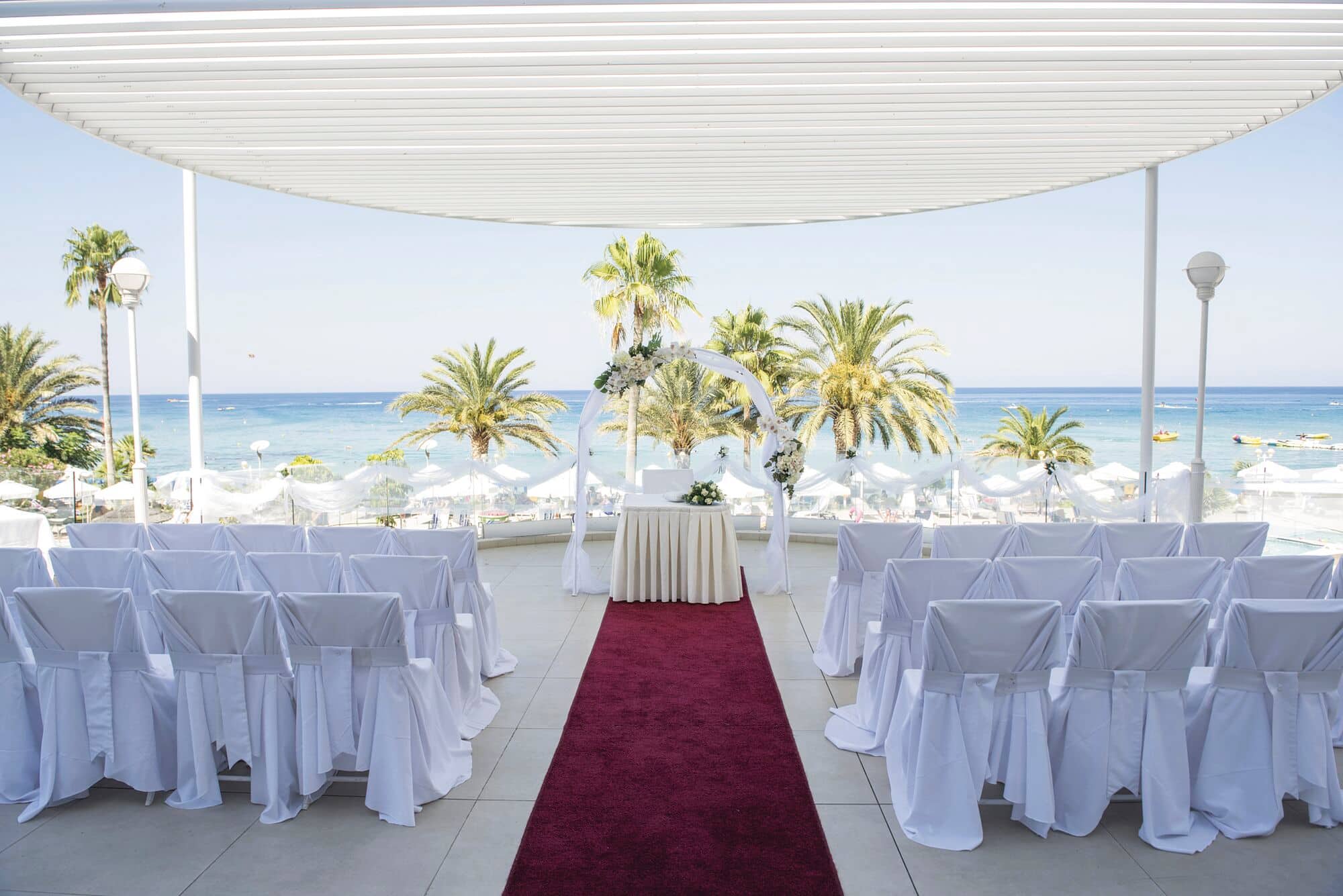 An elegant wedding ceremony sits under a shade, with a sunny beach scene in the background.