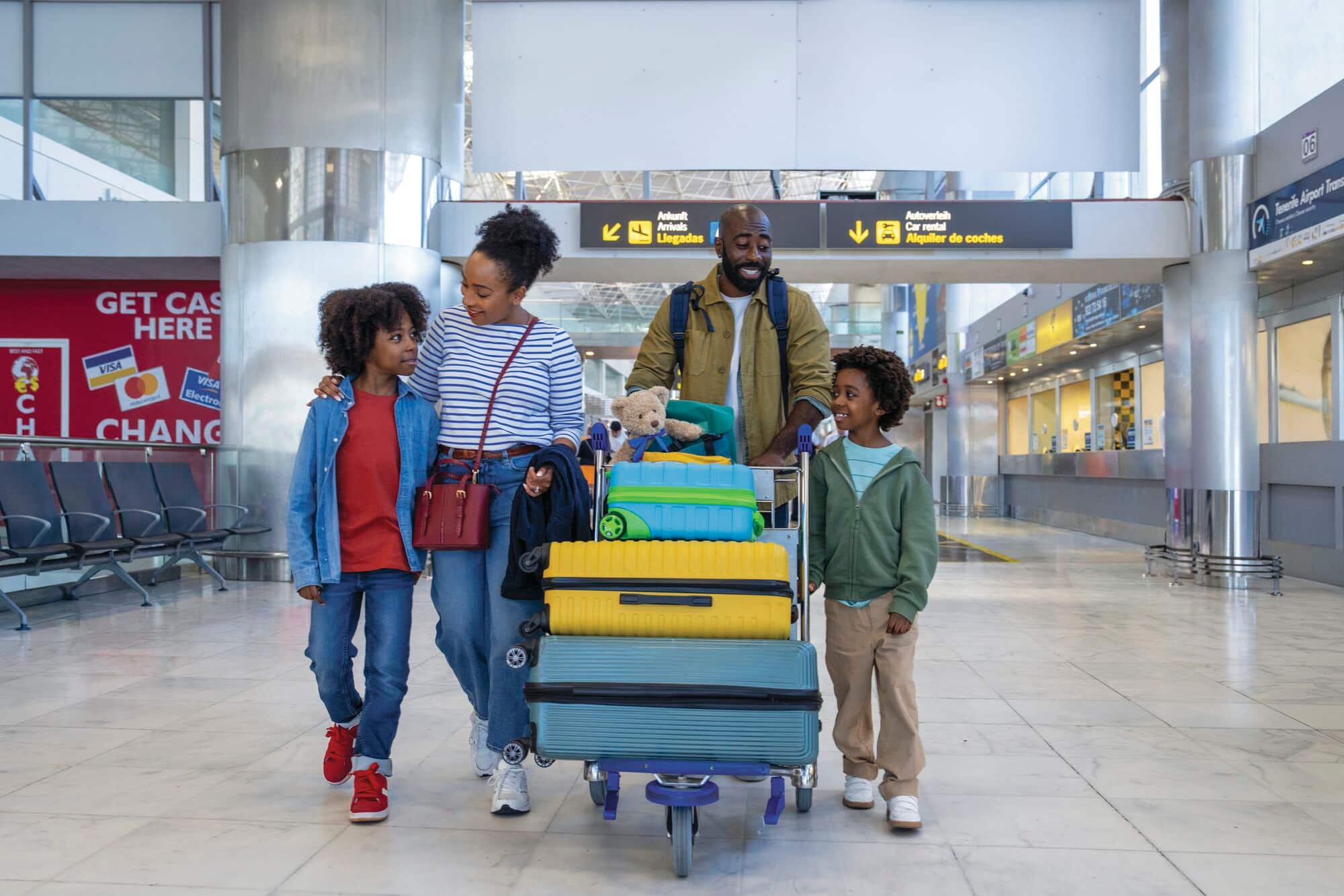 A family of four walking through an airport with a luggage trolley.
