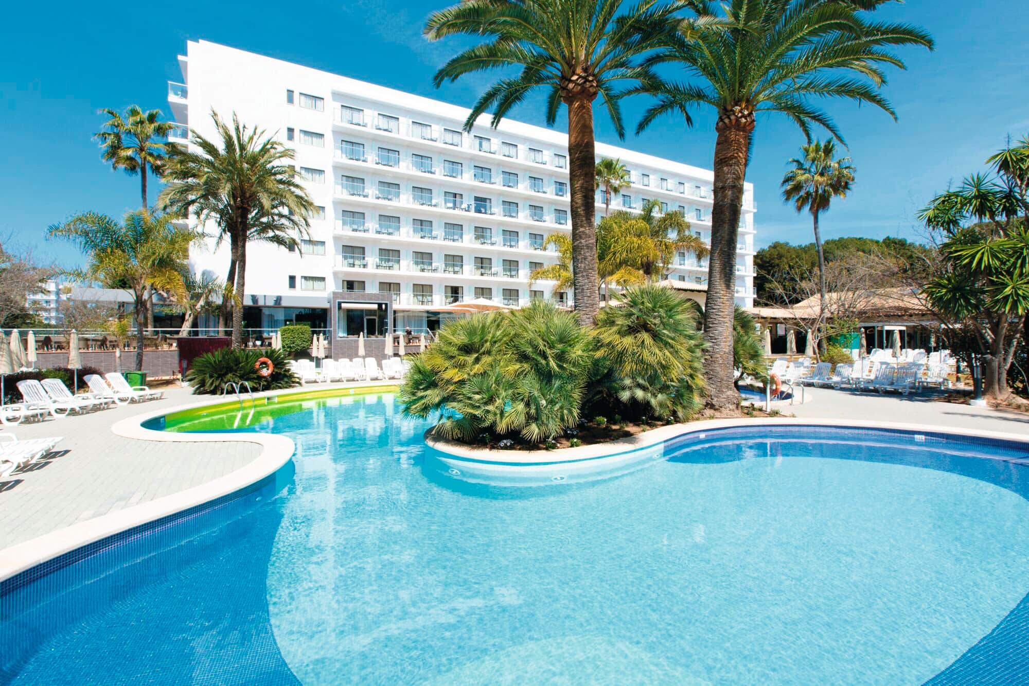 An outdoor swimming pool with curved edges surrounded by sun loungers and tall palm trees, in front of a modern white multi-story hotel building under a clear blue sky.
