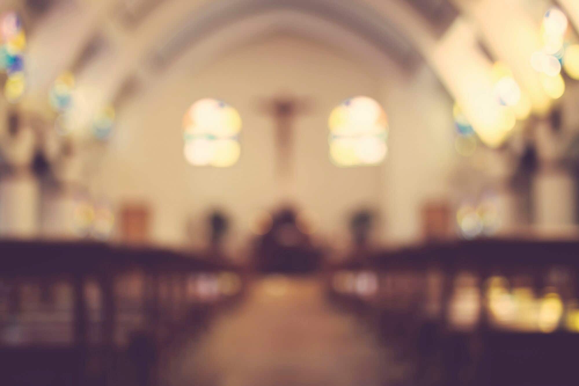 Blurred interior of a church with pews, stained-glass windows and a cross at the altar.