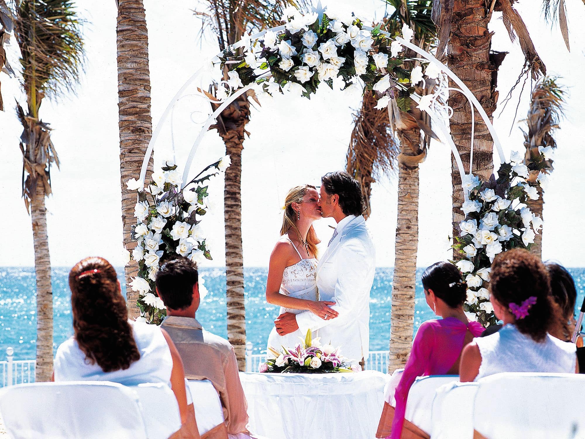 Bride and groom kiss in front of witnesses under a floral arch.