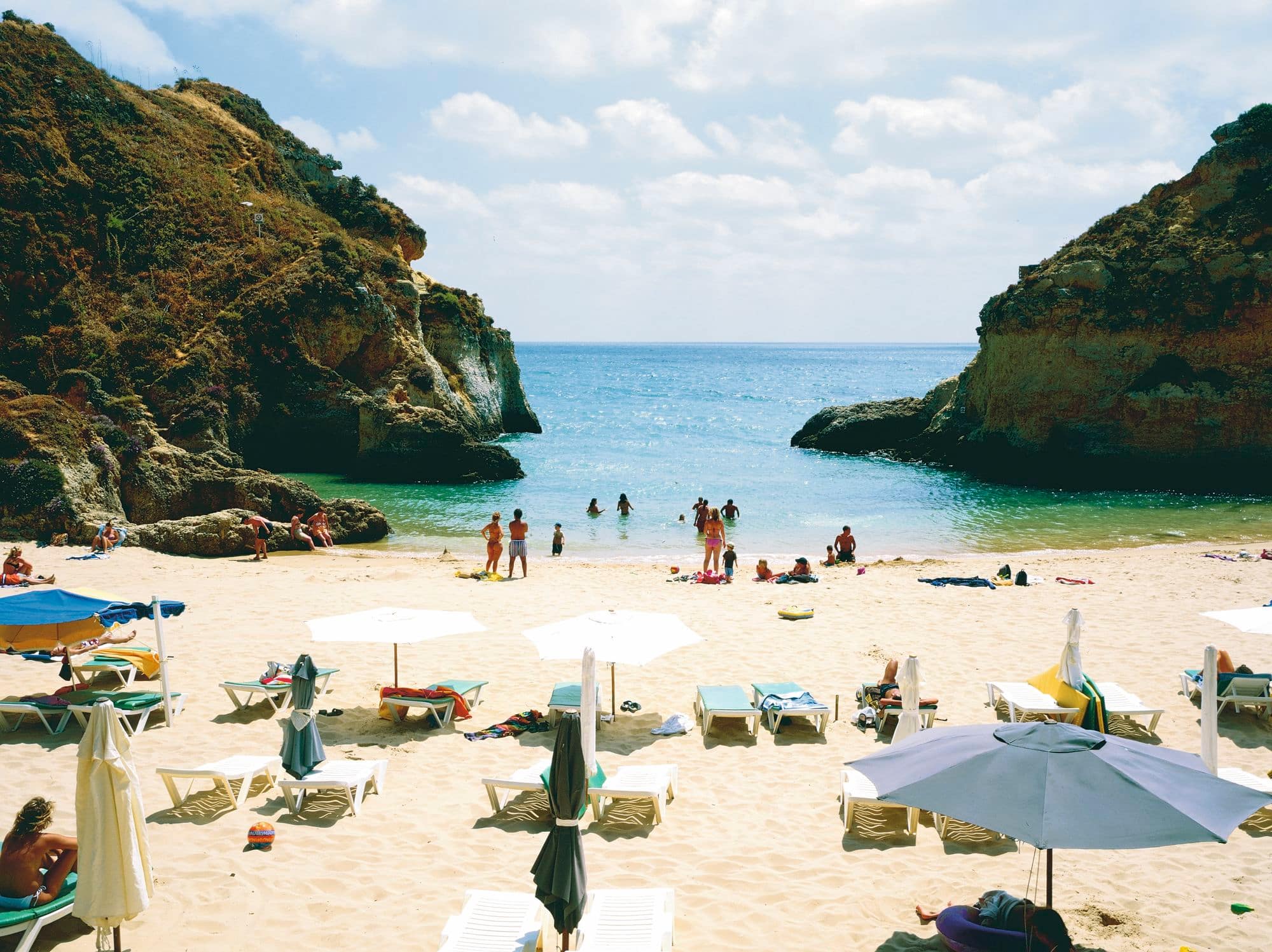 A beach cove surrounded by green rocky cliffs. White sun loungers and umbrellas are in the foreground, and swimmers are in the background.