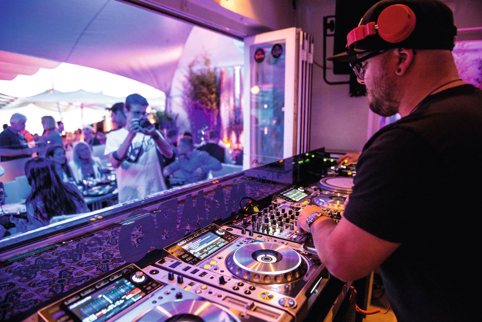A DJ wearing black cap and orange headphones performing on Pioneer equipment in a purple-lit booth.