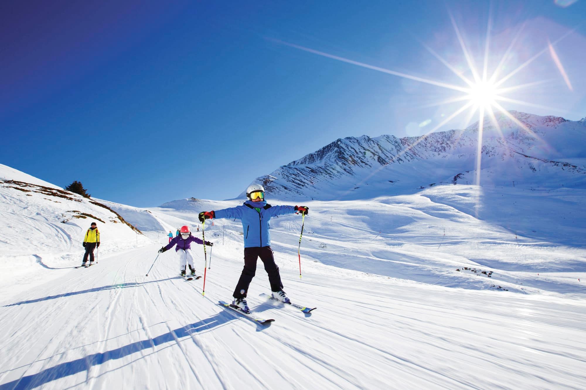 Three skiers travelling down a piste with arms wide under a blue sky and sunshine.