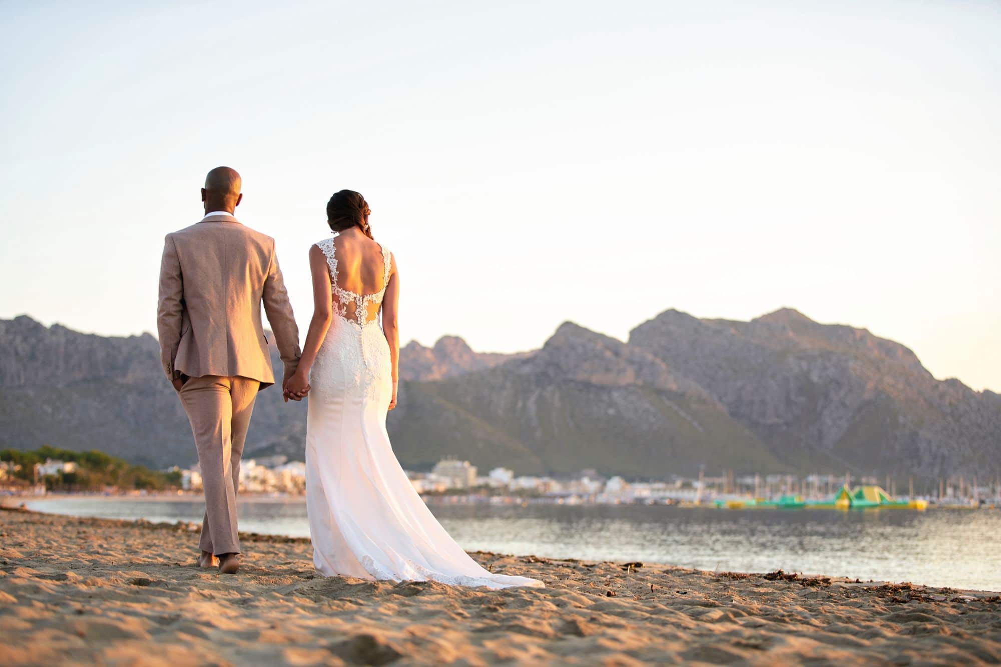 A bride and groom walk along a beach with mountains in the background.