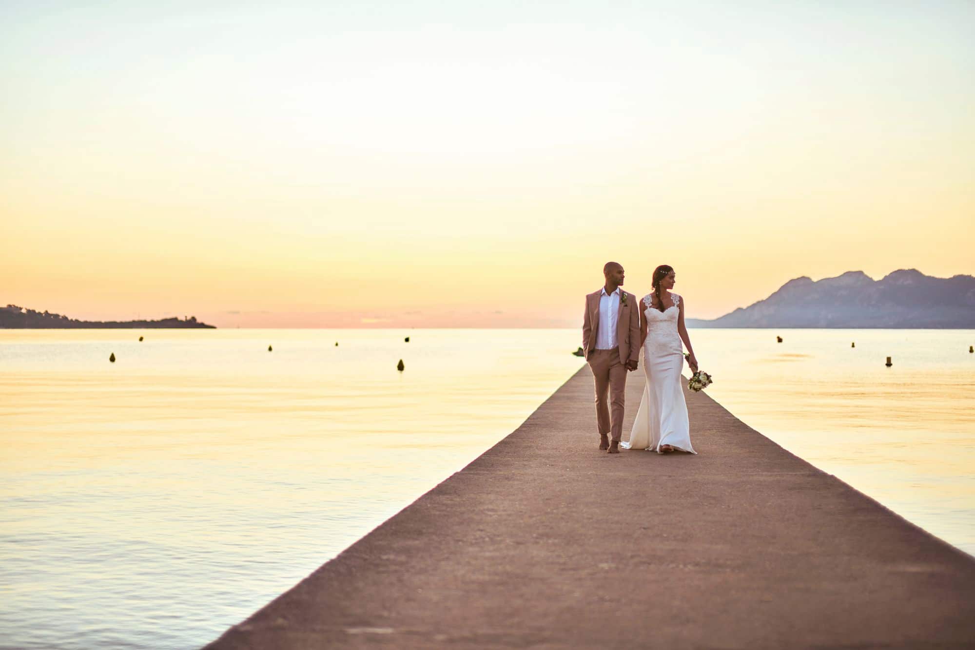 Bride and groom walking down a seaside promenade at sunset.