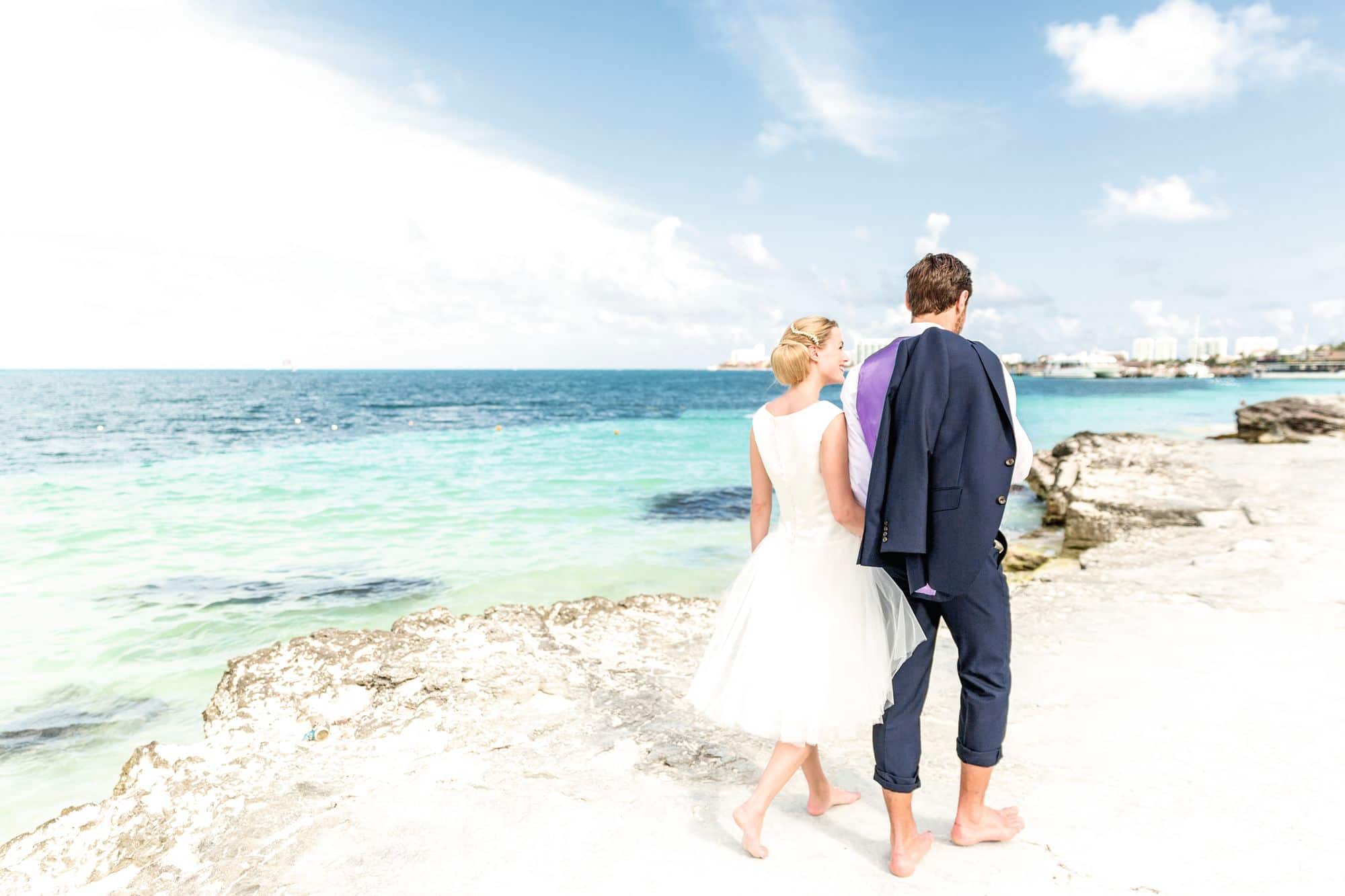 A couple on their wedding day standing barefoot on a white rocky coastal shoreline.