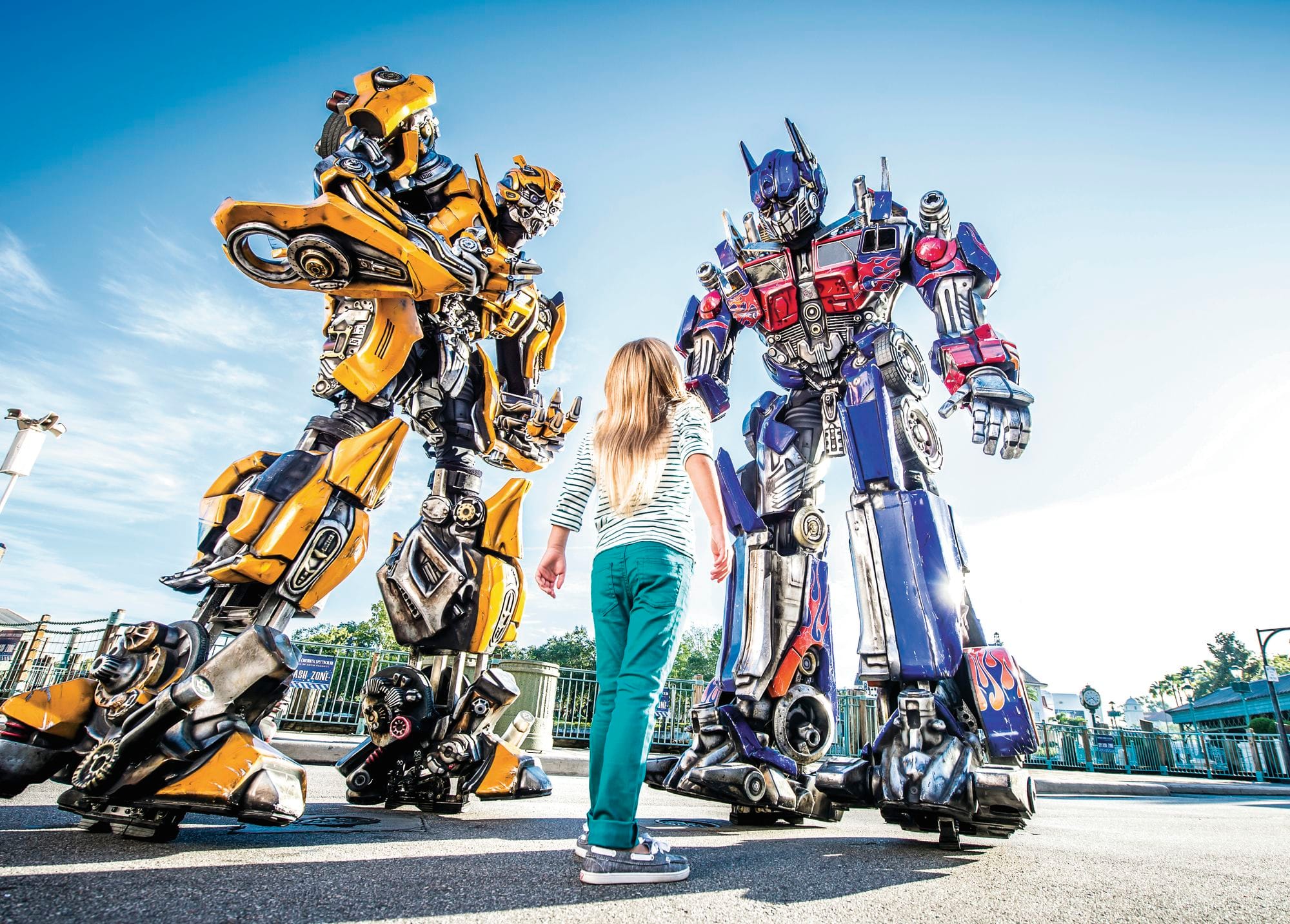 A little adventurer meets a pair of giant Transformers robots under a clear sky.