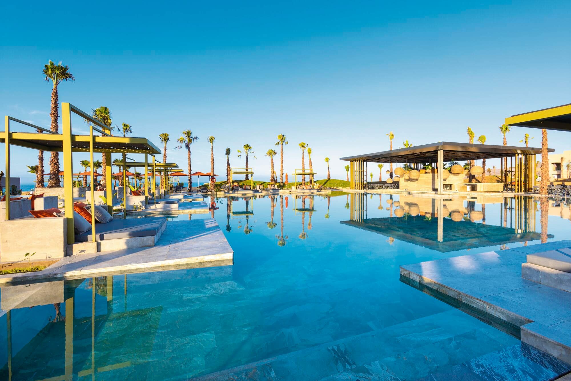 Luxury resort infinity pool with turquoise water, surrounded by yellow-roofed pavilions and palm trees.