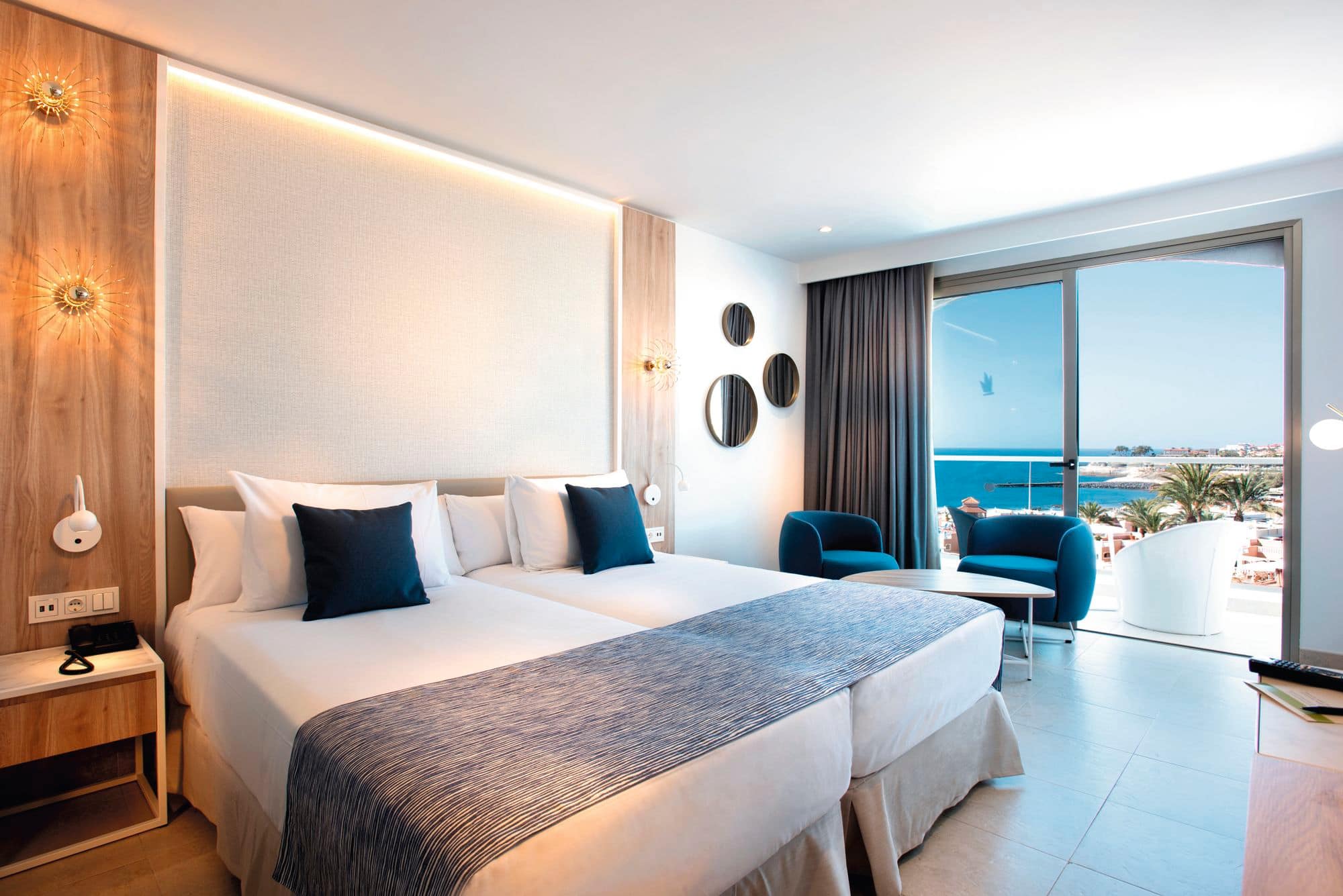  A hotel room with a view of the sea. The beds are decorated with blue runners and pillows.