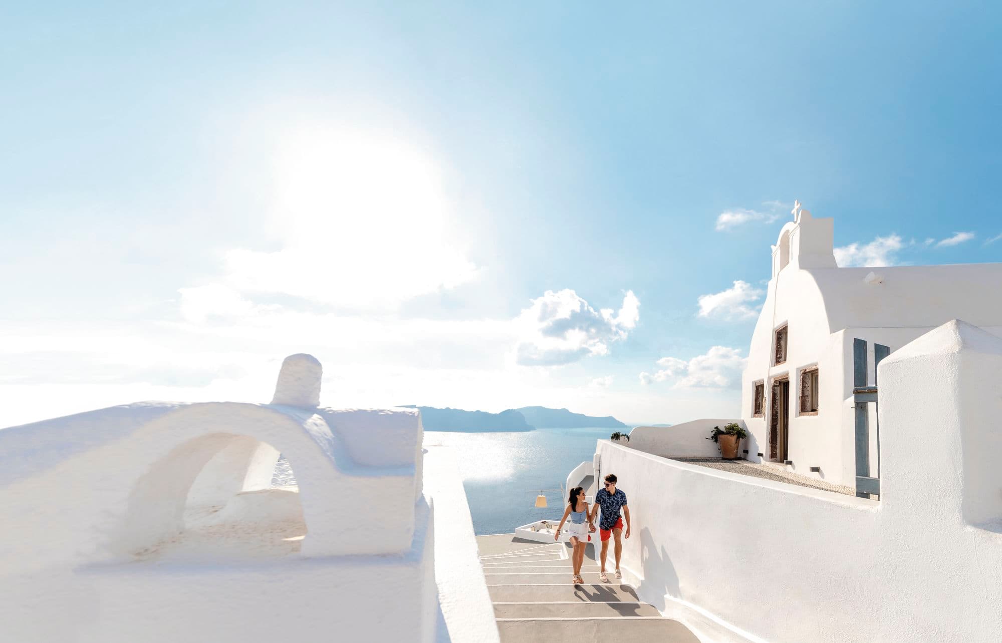 A couple walking up whitewashed steps between traditional Cycladic-style buildings.