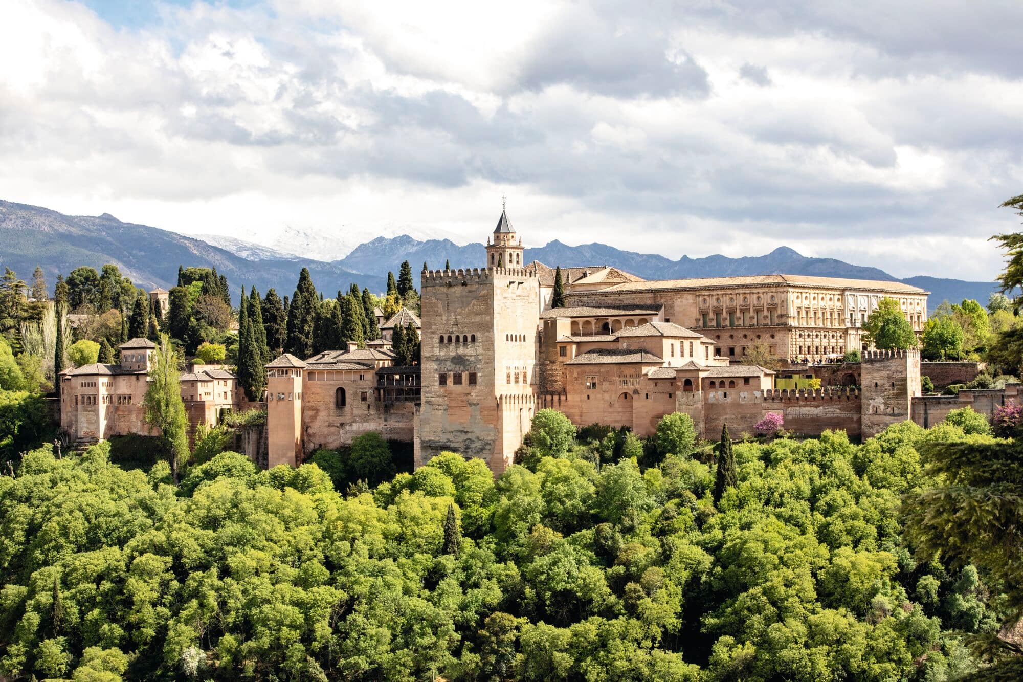 The Alhambra in Granada, overlooking green woodland with mountains behind.