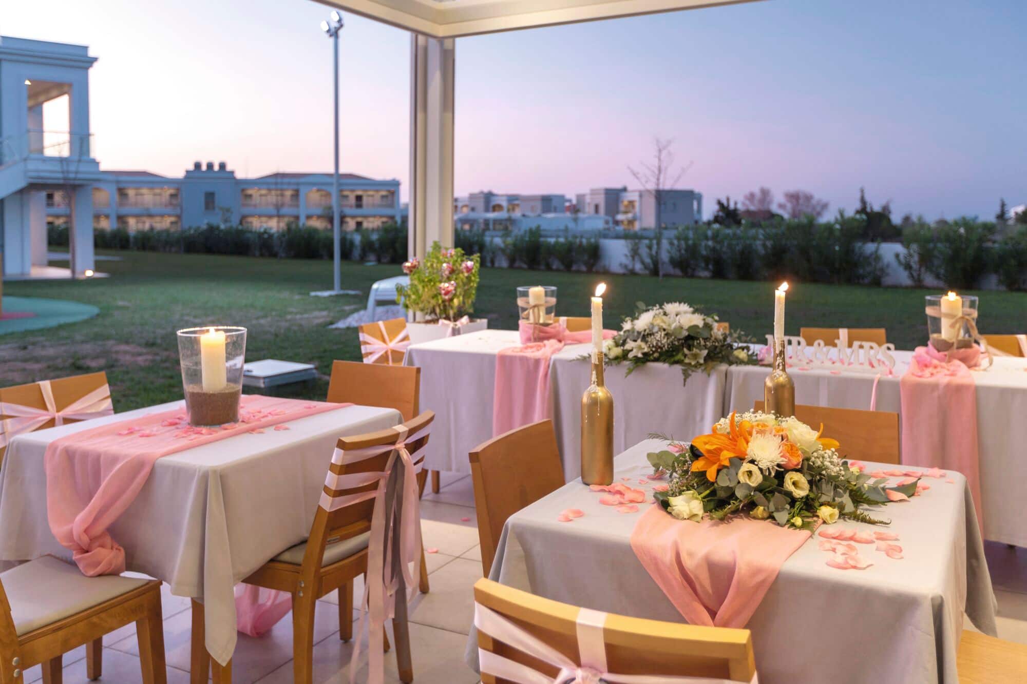 An outdoor wedding dining area, with tables decorated with pink runners, floral centrepieces, candles, and scattered rose petals.
