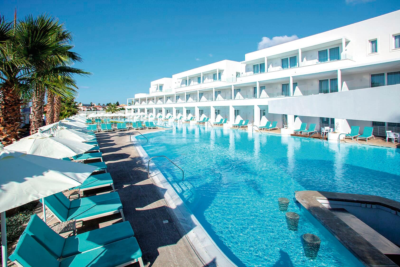 Long outdoor swimming pool lined with turquoise sun loungers and white umbrellas, adjacent to a modern white multi-story hotel building with balconies. Palm trees border the pool area under a bright blue sky.