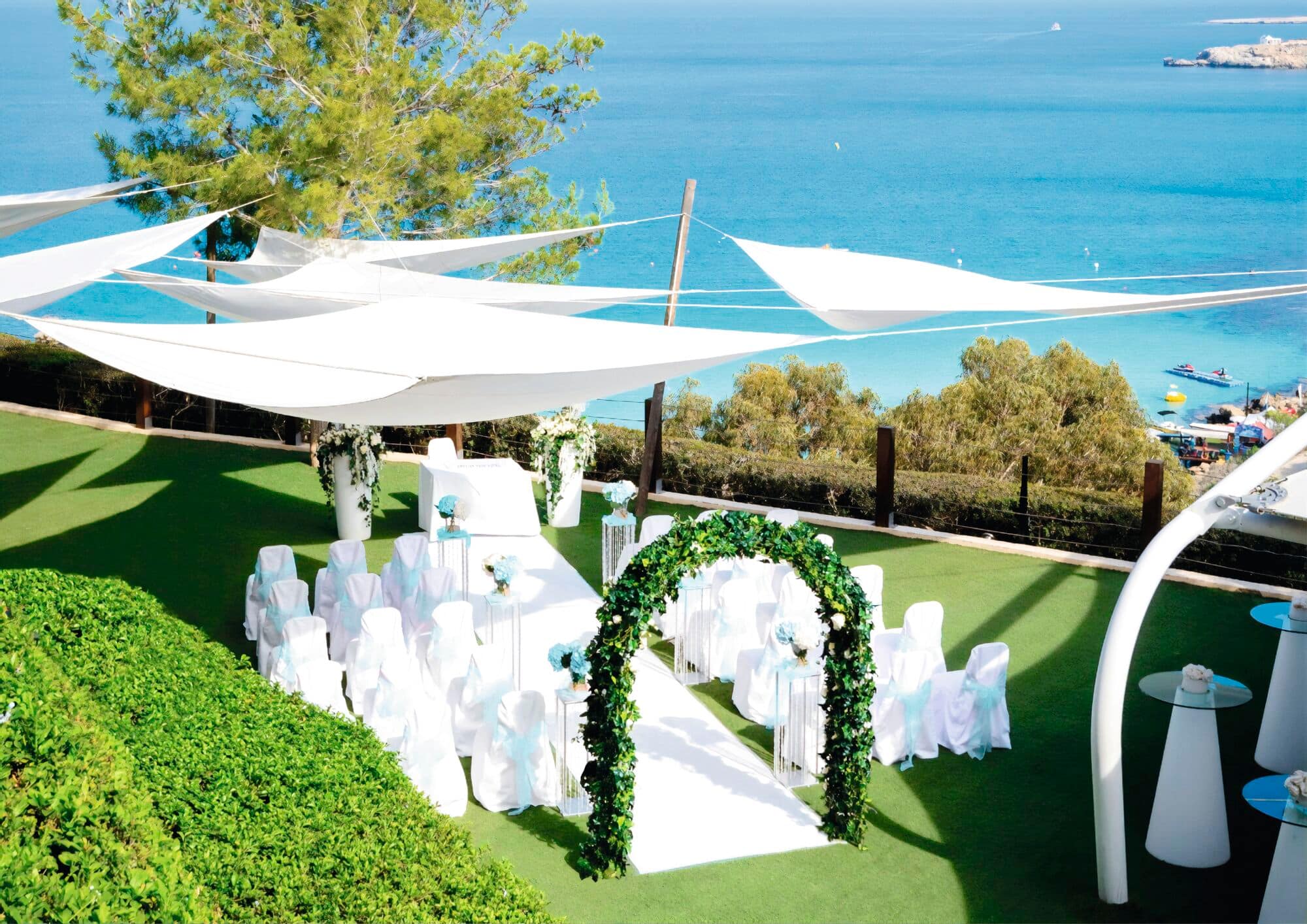 An outdoor wedding setup on a green lawn overlooking a bright blue sea.
