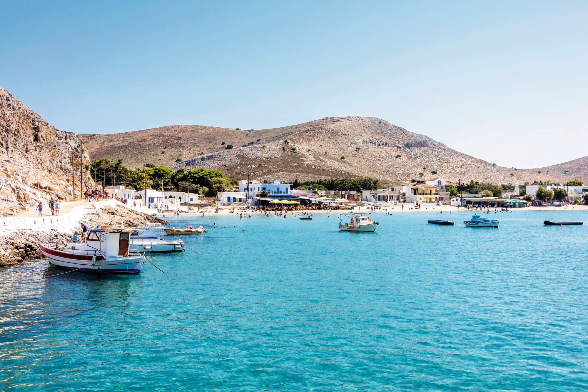 Mediterranean coastal village with white buildings nestled at the base of arid tan hills, overlooking crystal-clear turquoise water with several small boats anchored in the bay.