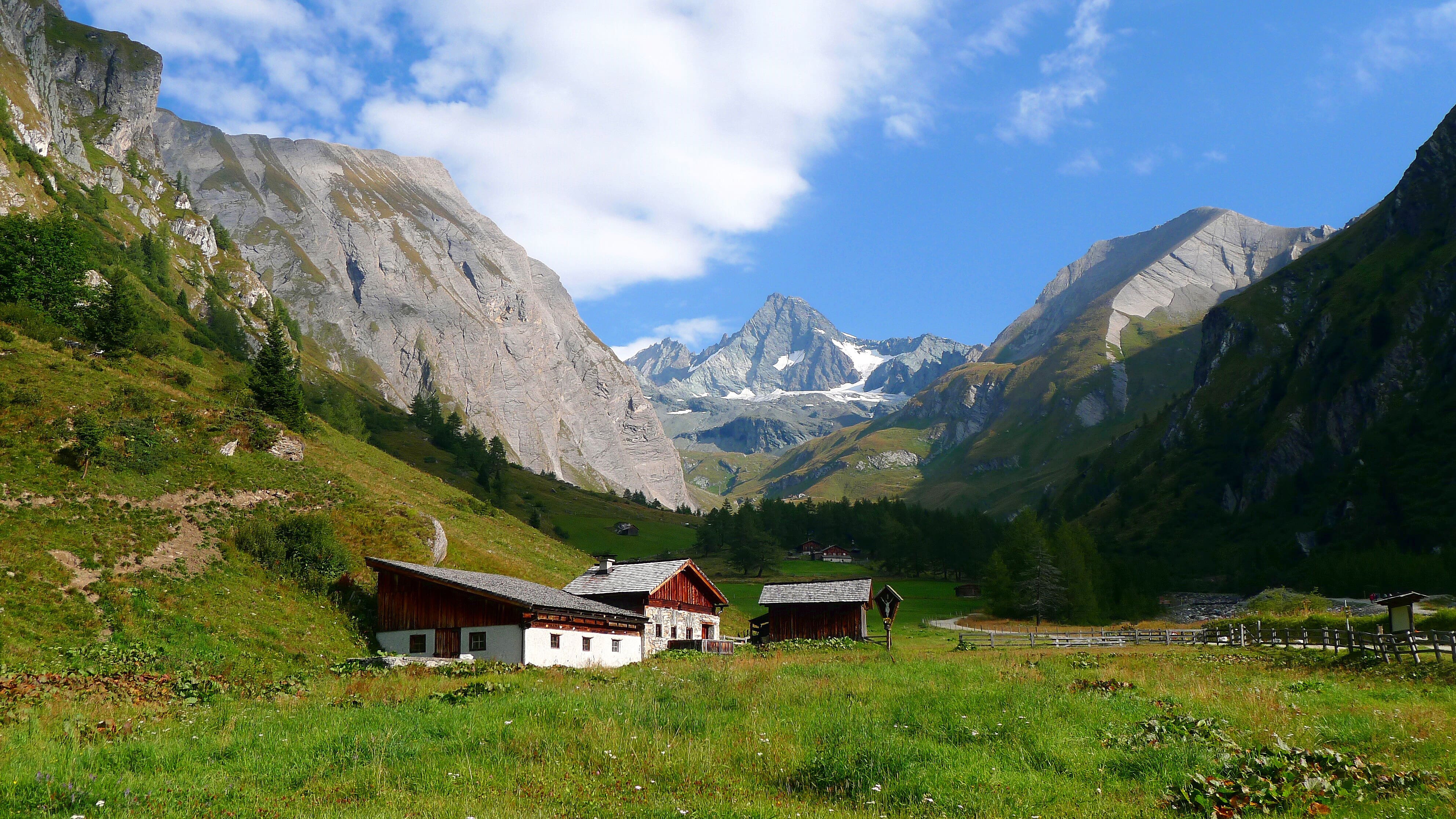 Alpine valley with three small huts - a pointed rocky peak is in the distance. 