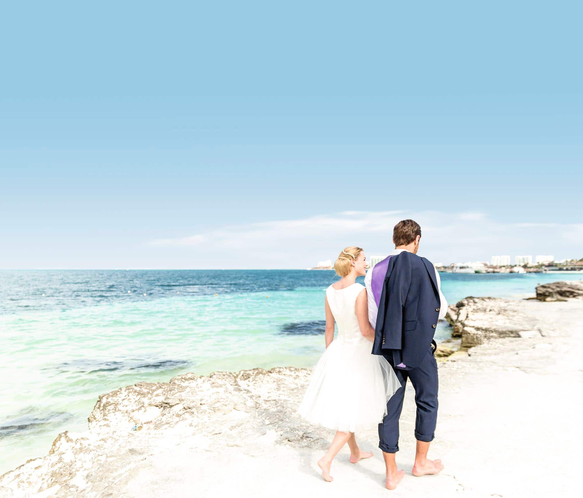 A bride and groom walk barefoot by the sea.