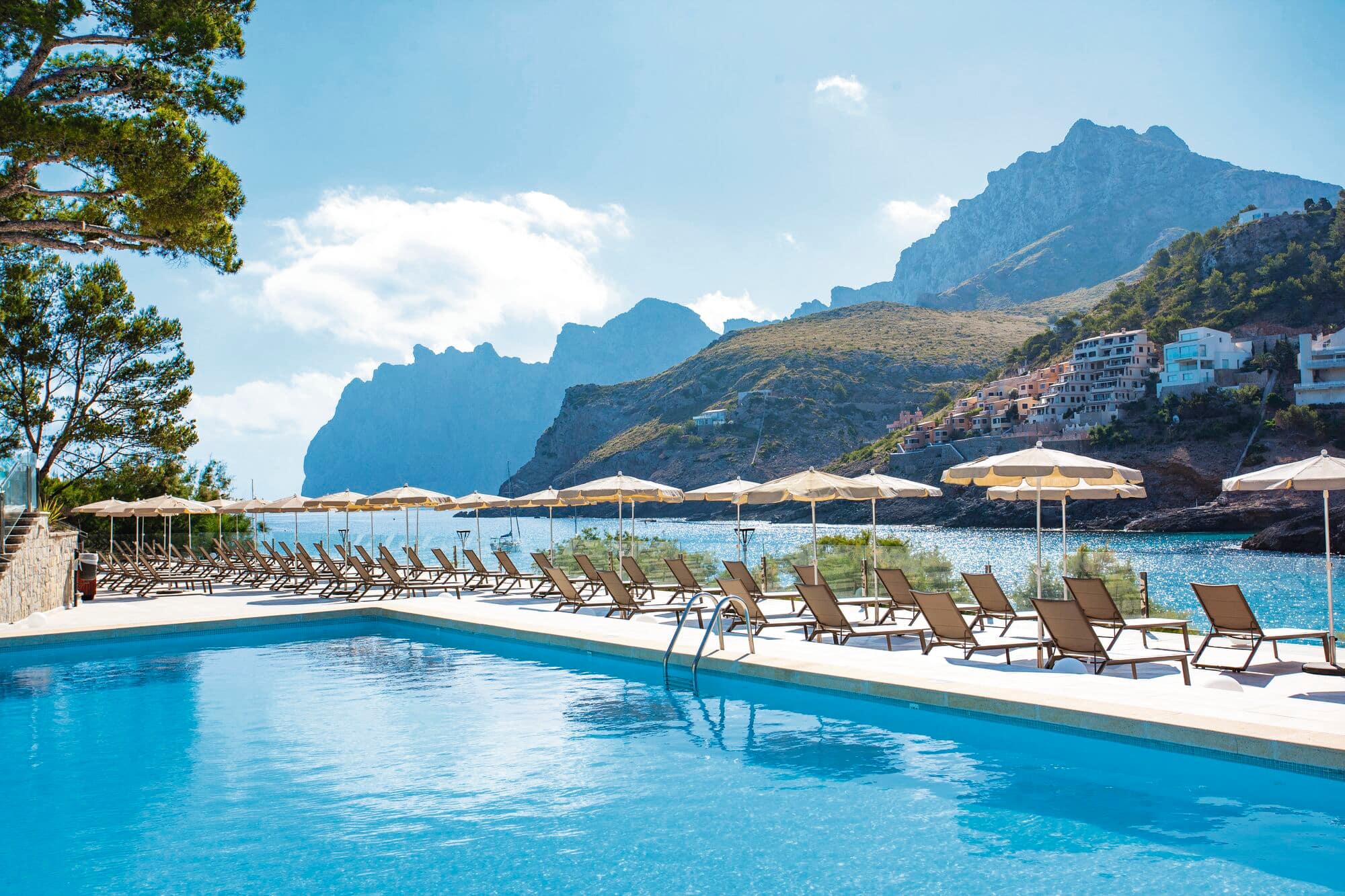 Outdoor swimming pool with rows of sun loungers and umbrellas overlooking a scenic bay.