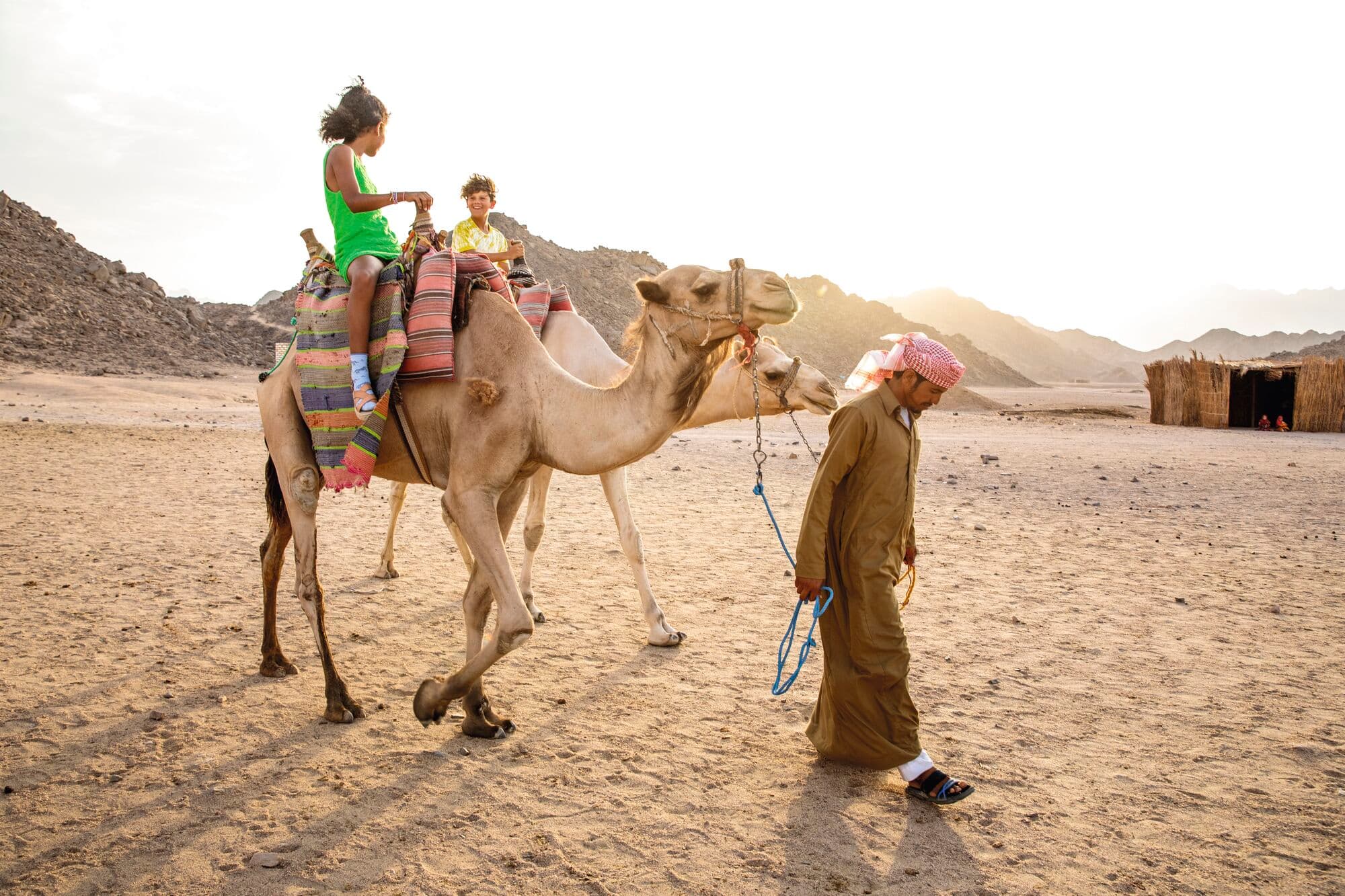 A man in traditional Middle Eastern dress leading a camel with  colourful striped saddle blanket carrying a woman and child through the desert.