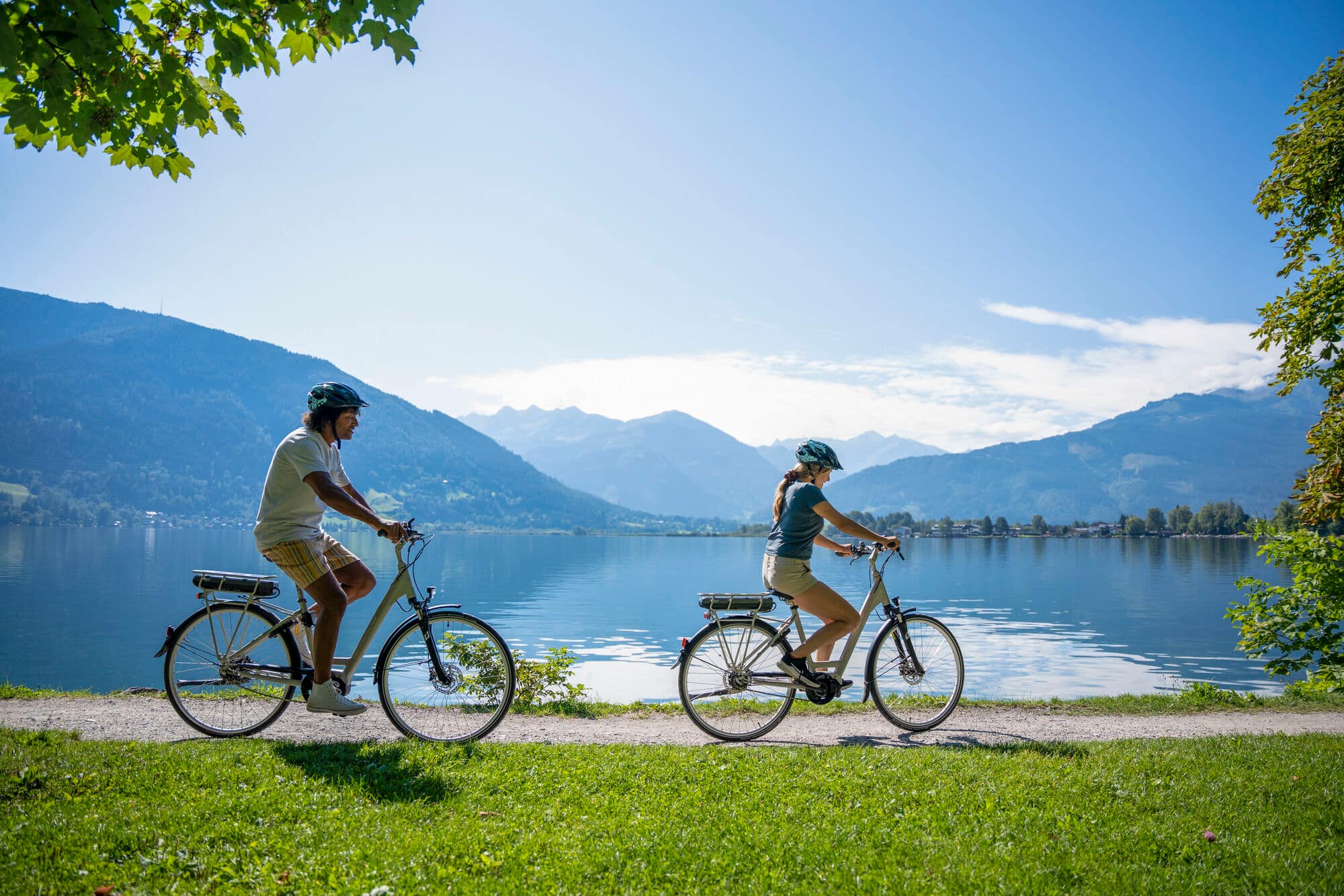 A man and woman cycling behind one another along a flat path beside a lake. 