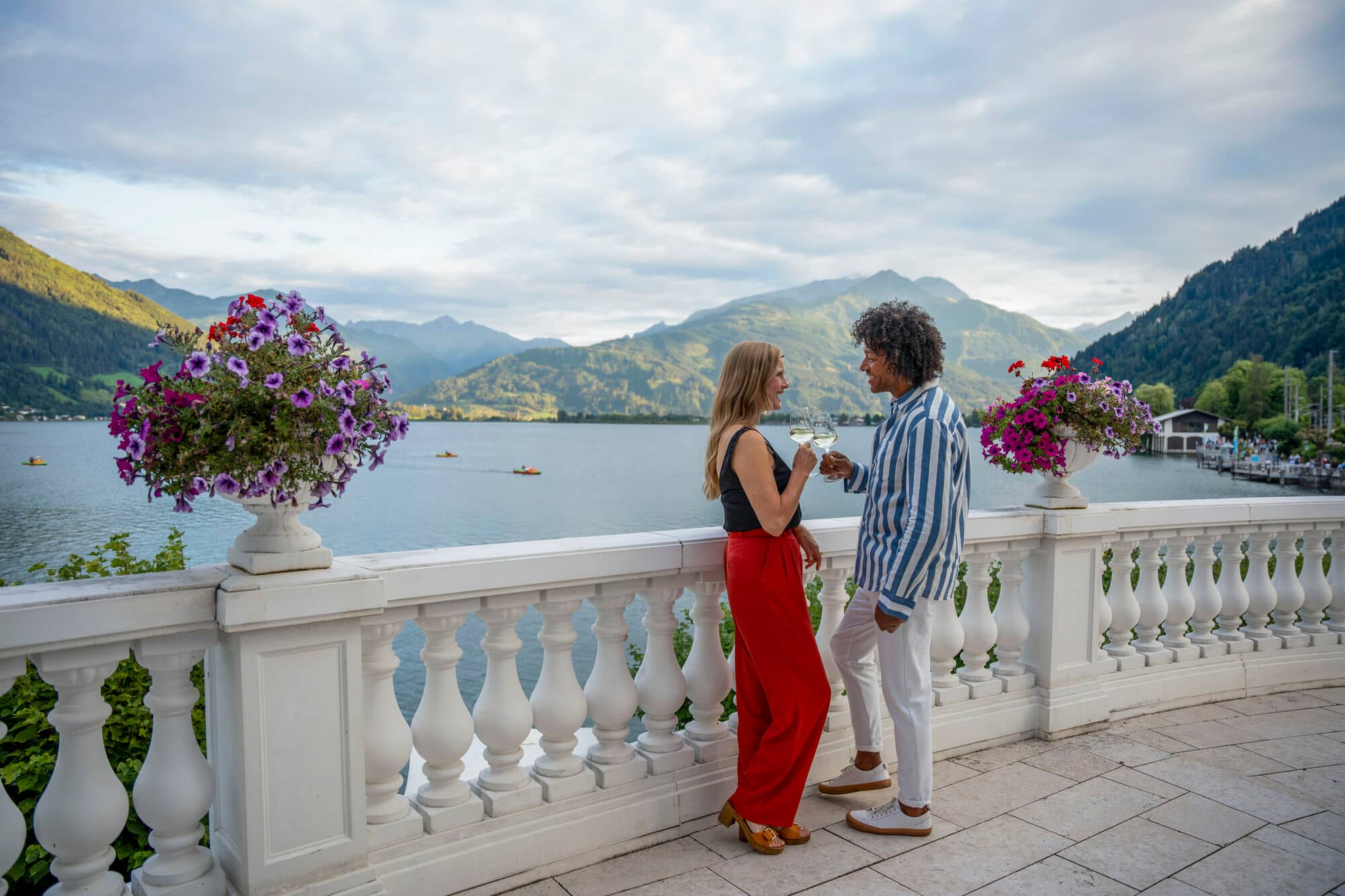 Couple enjoying wine on a balcony overlooking a serene lake and mountain landscape, with purple petunias blooming in planters along the white railing.