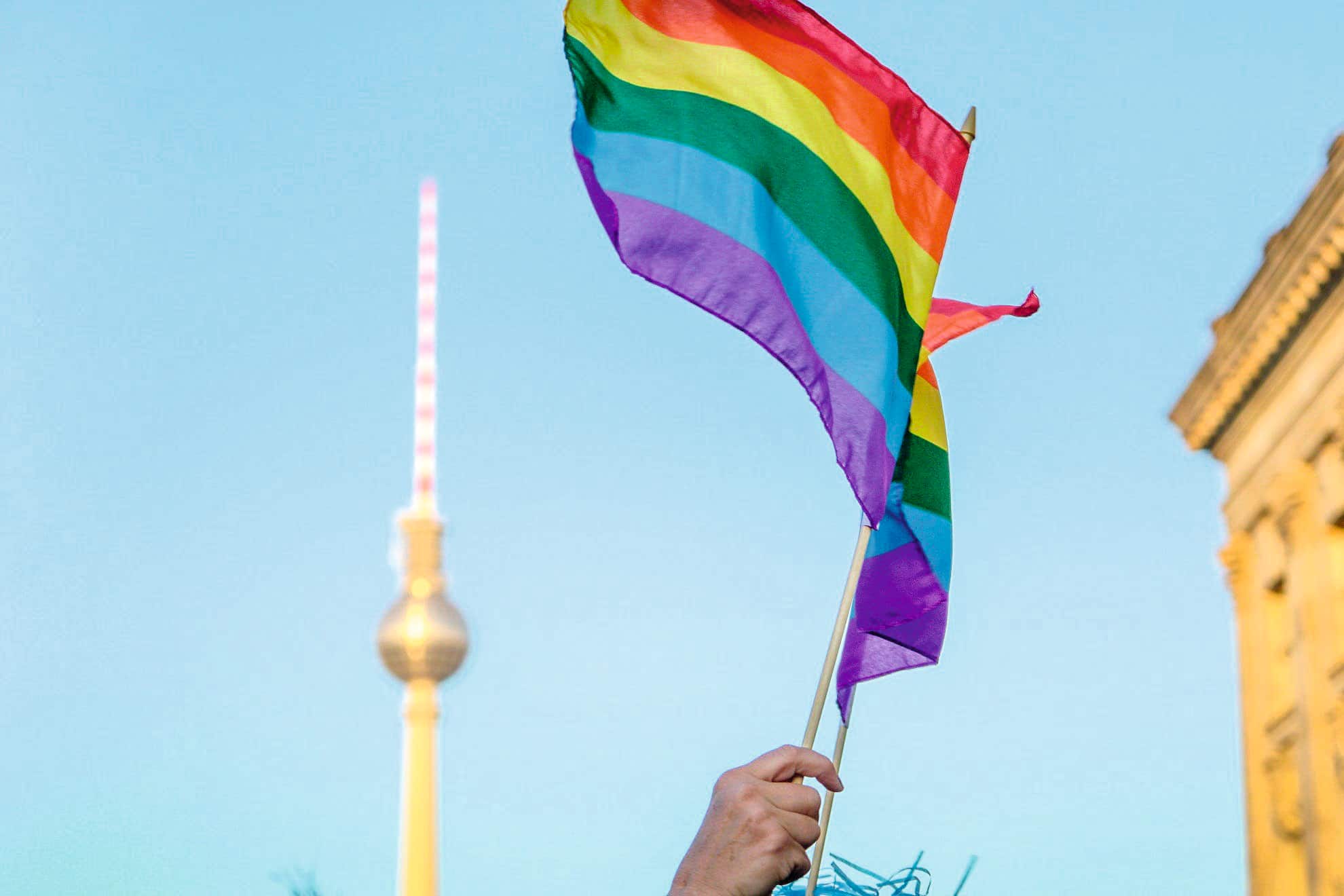 A hand holds a rainbow Pride flag waving against a clear blue sky.