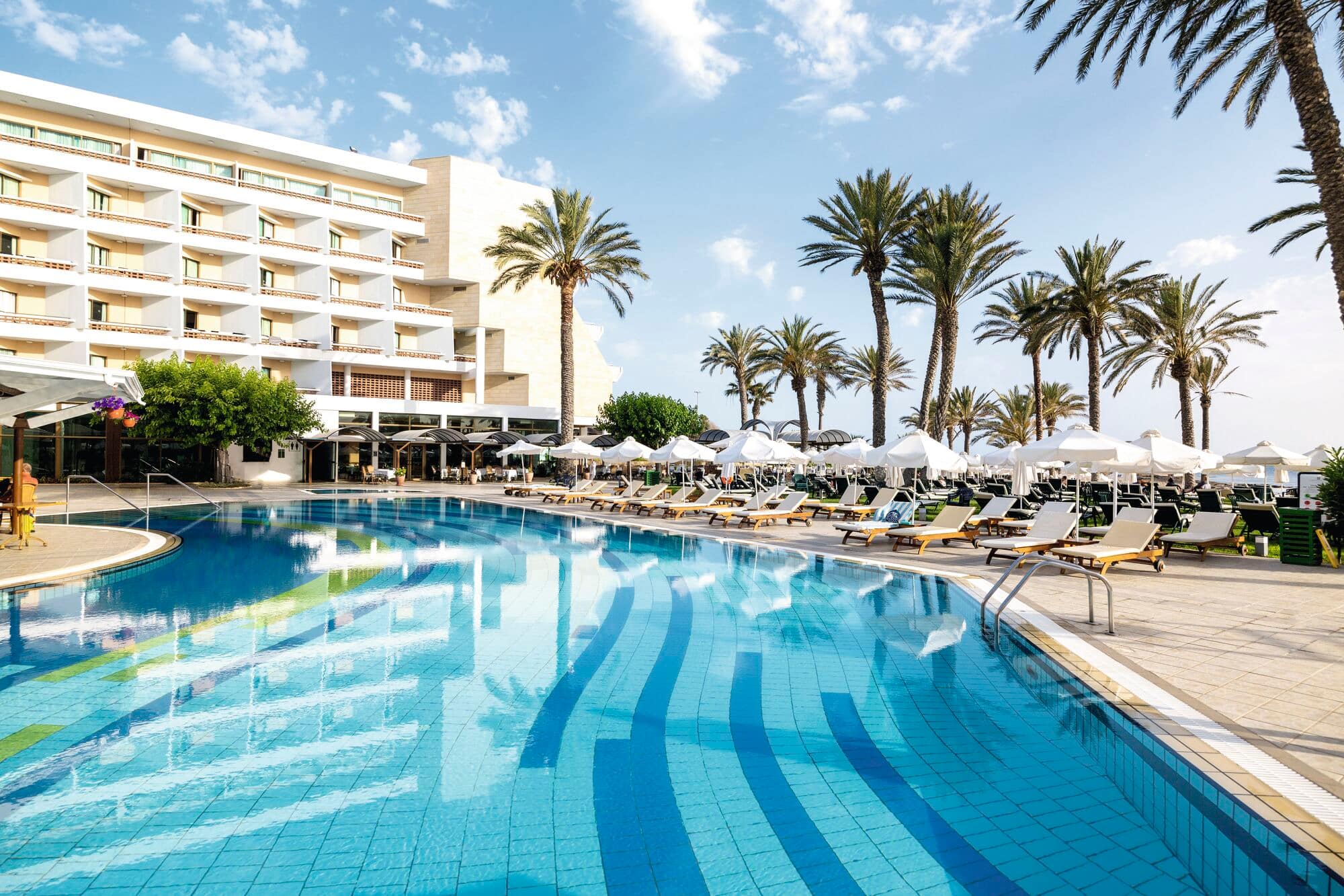 Outdoor swimming pool with clear blue water surrounded by sun loungers and white umbrellas, set against a backdrop of tall palm trees and a multi-story hotel building under a partly cloudy sky.