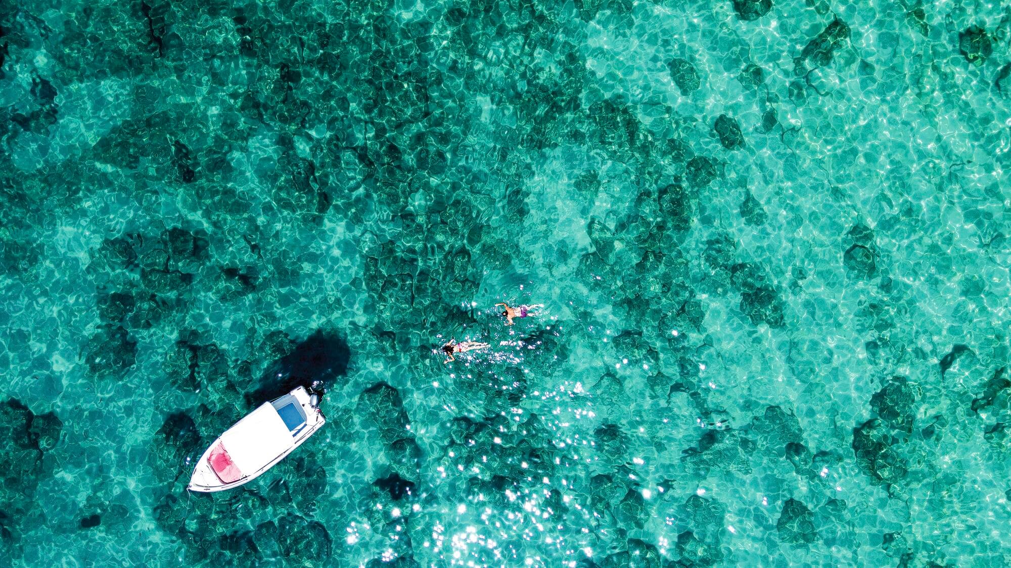 Overhead view of two people swimming by a boat in a clear blue sea.