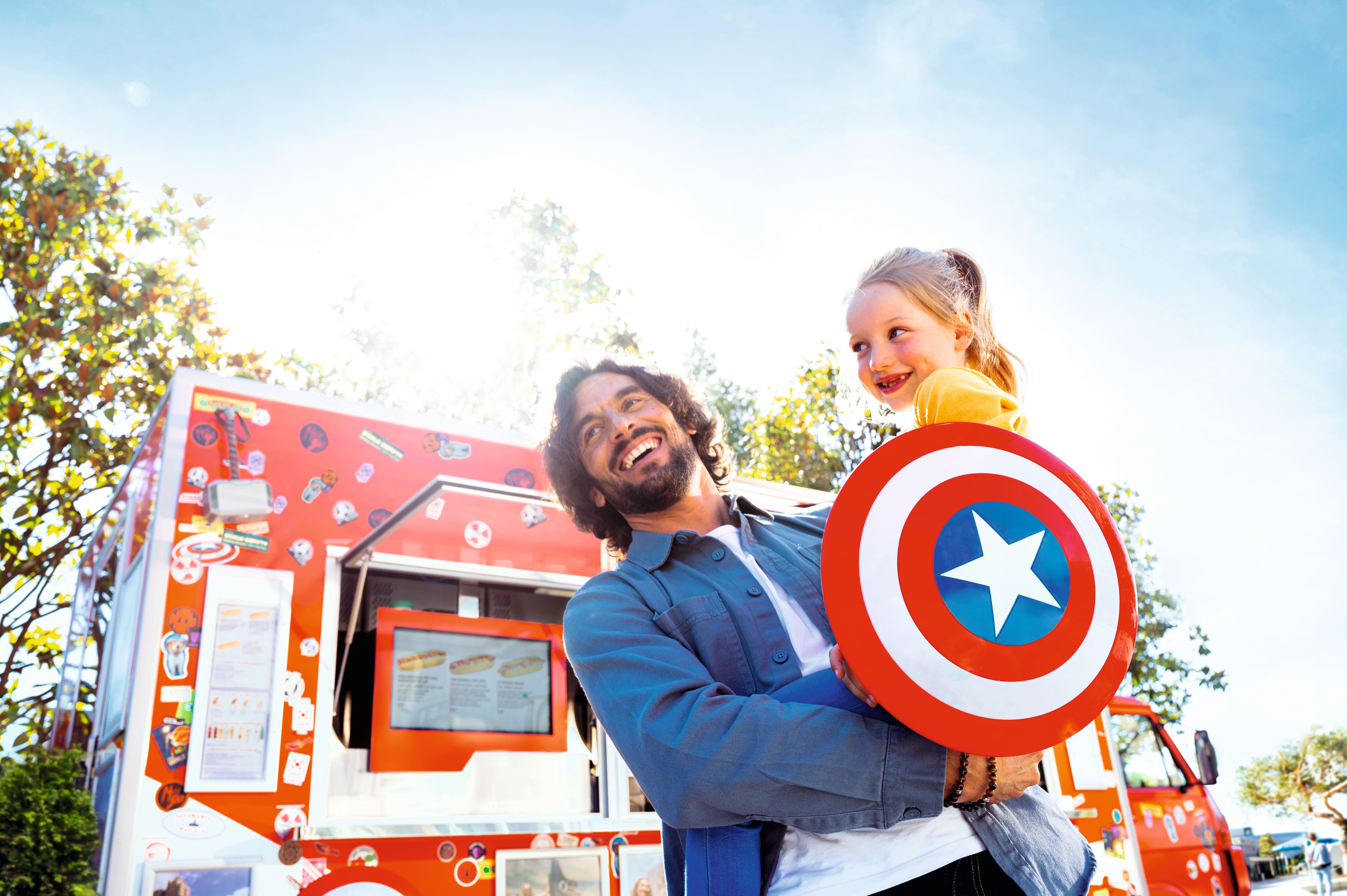 A parent and child holding a Captain America shield in front of a food truck.