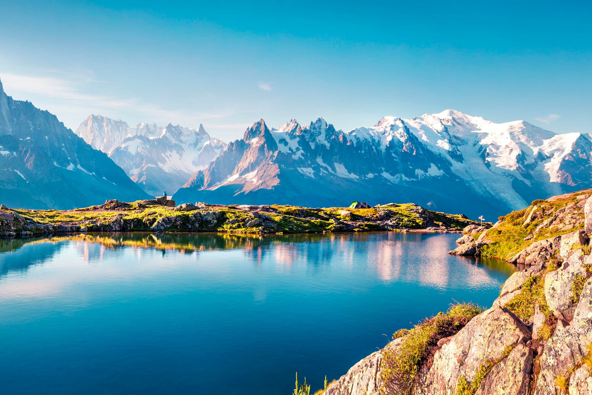 A clear blue lake, with Mont Blanc and surrounding mountains reflected in it.