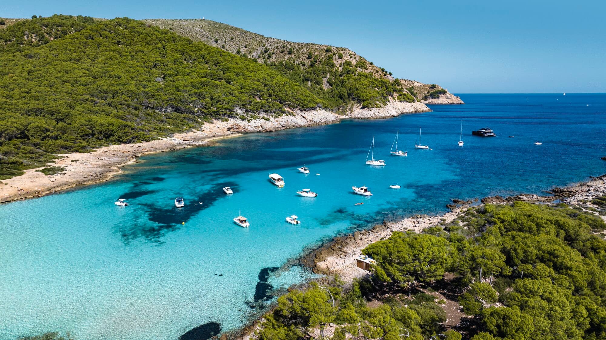 An aerial view of a turquoise bay in Majorca, surrounded by pine-covered hills and the Mediterranean Sea.