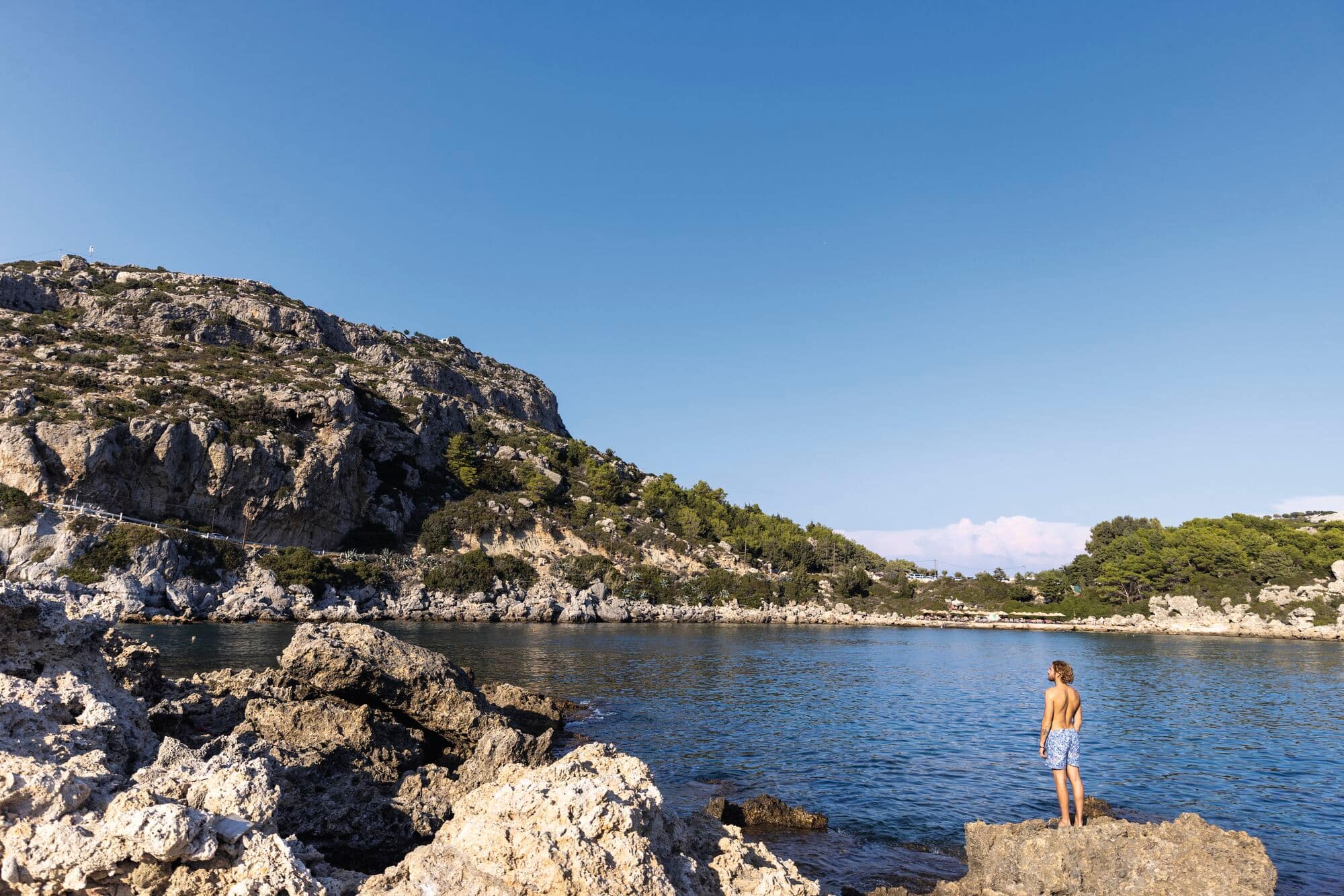 A person stands on limestone rocks overlooking a calm, deep blue cove, with a dramatic beige cliff face on the left.