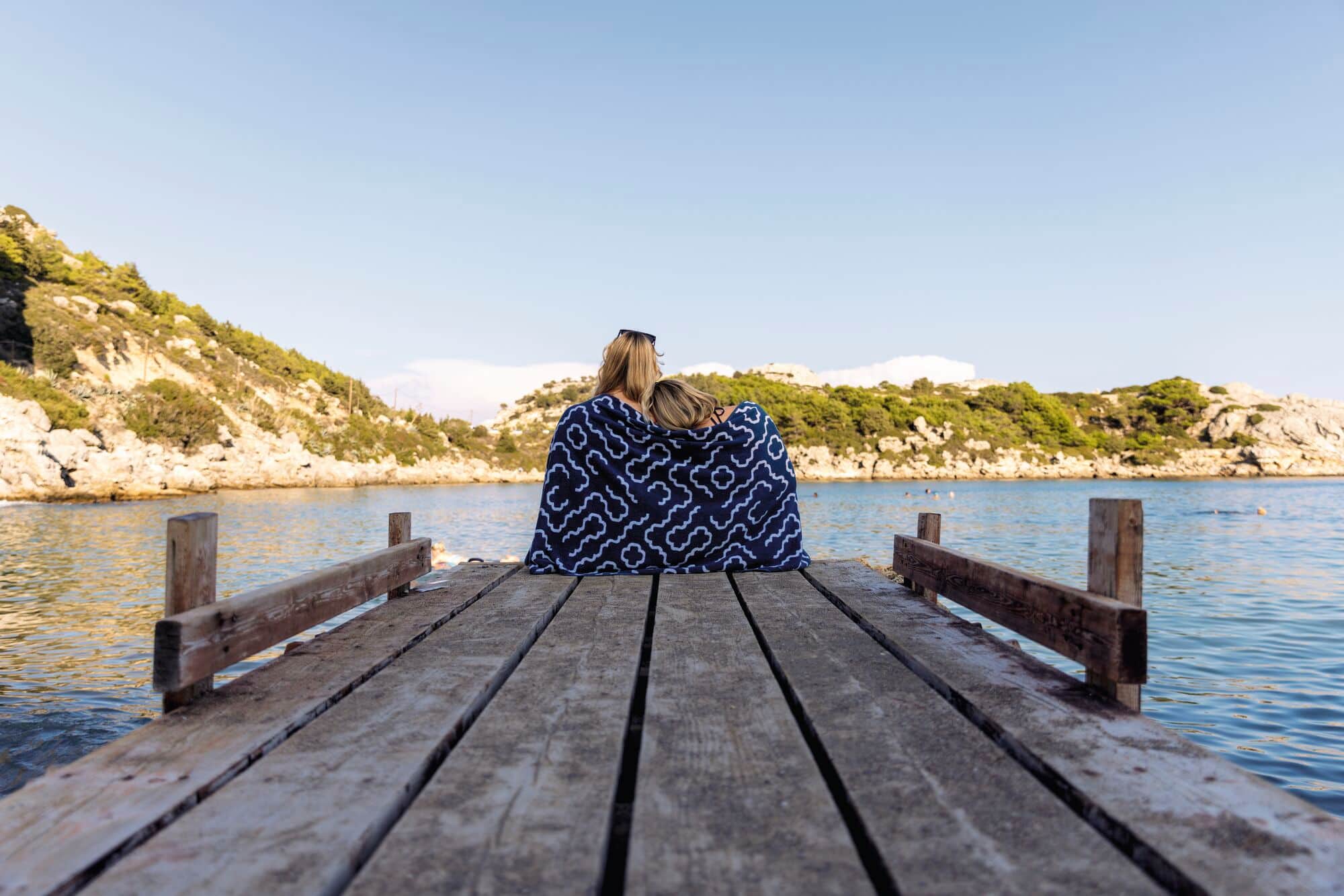 Two people sat together on a jetty with a towel over their shoulders.