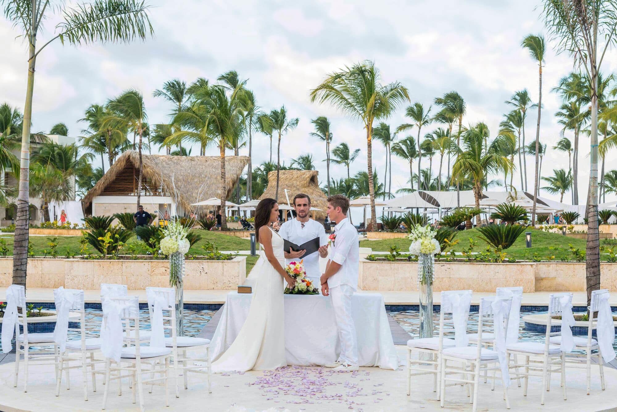 A couple in white stand before an officiant by the hotel pool, surrounded by palm trees.