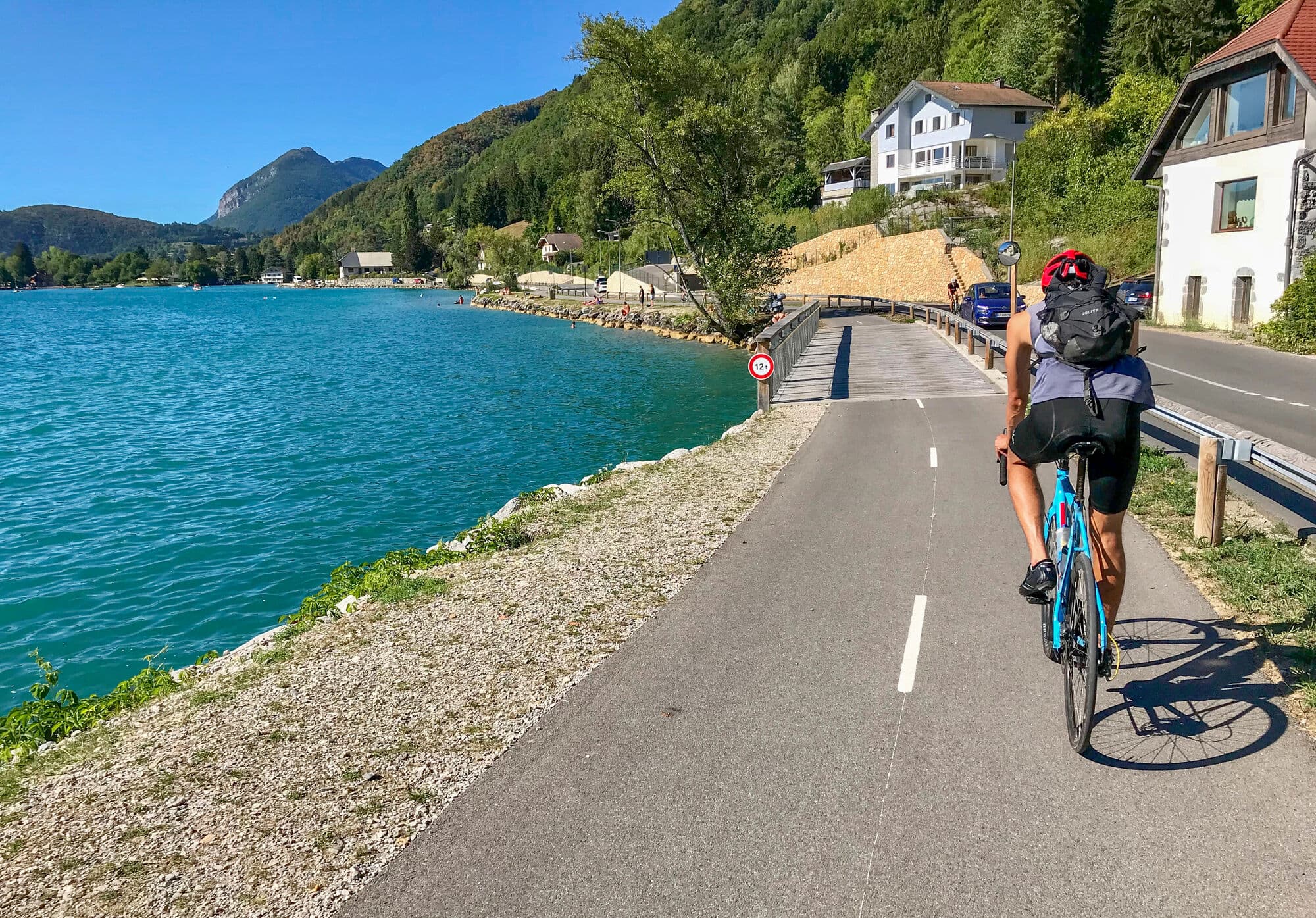 A cyclist with a backpack on seen cycling from behind on a tarmac path alongside a lake. 