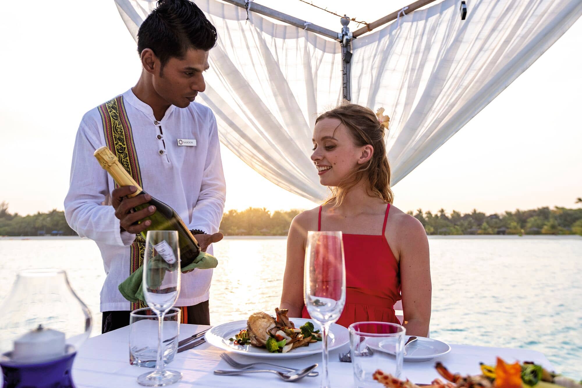 A waiter holding a champagne bottle at a seaside dining table that's set up with a white tablecloth, gourmet meal and wine glasses.