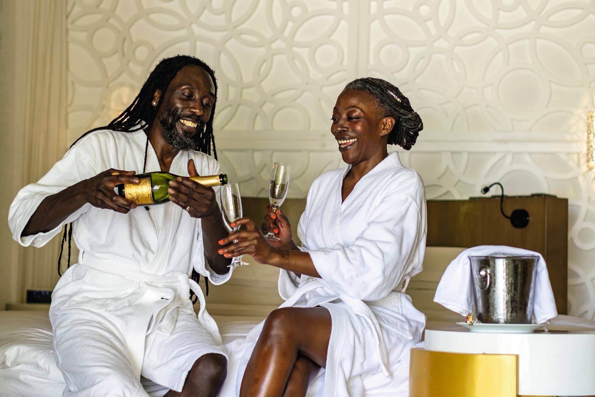 A couple in white bathrobes celebrating with champagne in a hotel room.