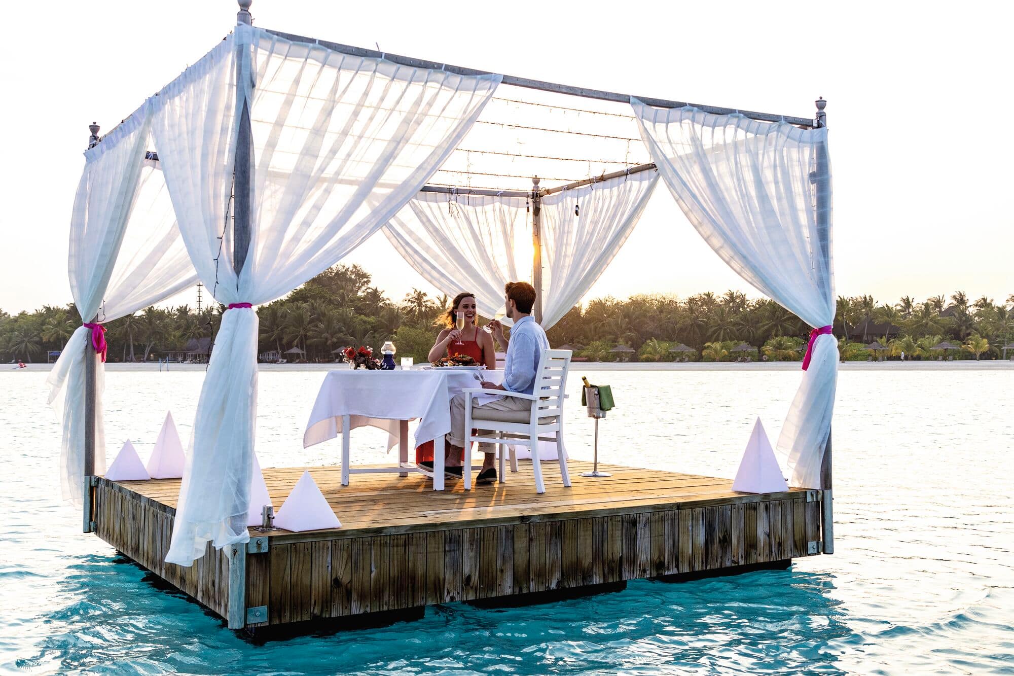 Romantic floating dining platform on turquoise water with white canopy, two diners at a table, and tropical shoreline in the background at golden hour.