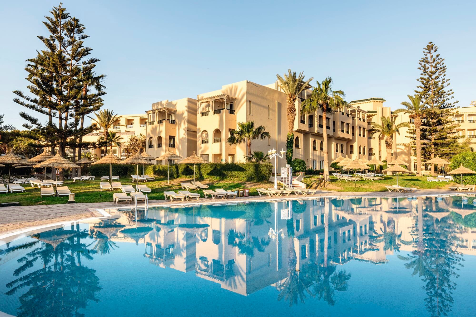 A blue swimming pool surrounded by sun loungers and palm trees