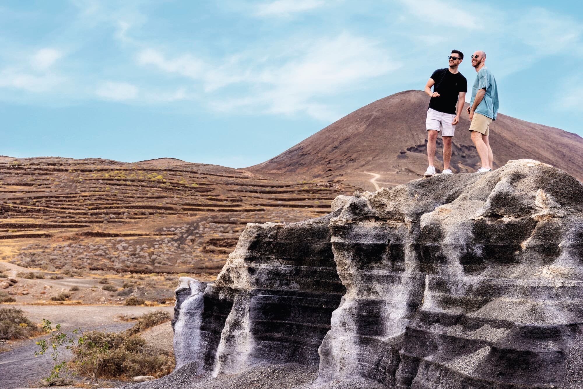 Two men standing on a layered rock formation overlooking a volcanic landscape.