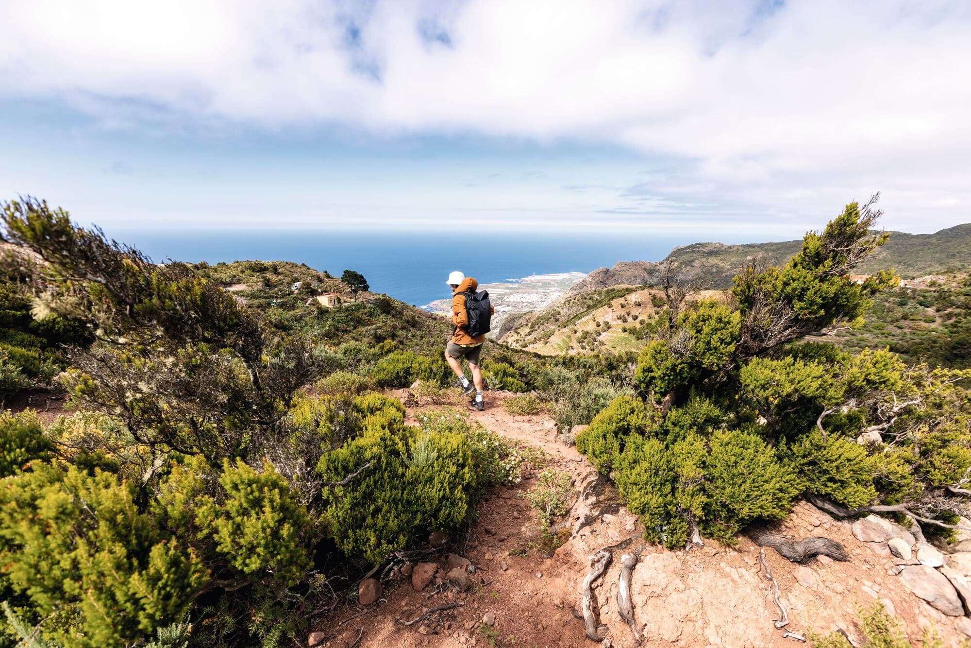 A hiker with an orange backpack walks down a rocky coastal trail.