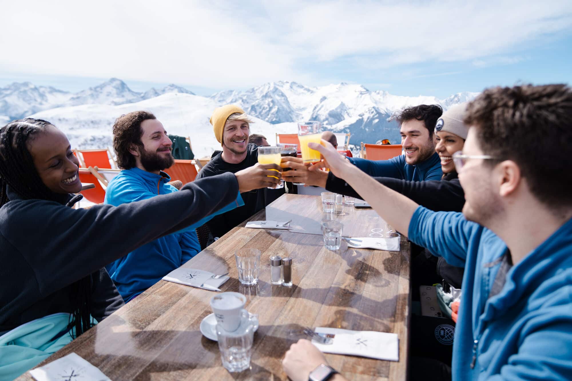 A group of people in ski gear clinking drinks at a table overlooking the mountains. 
