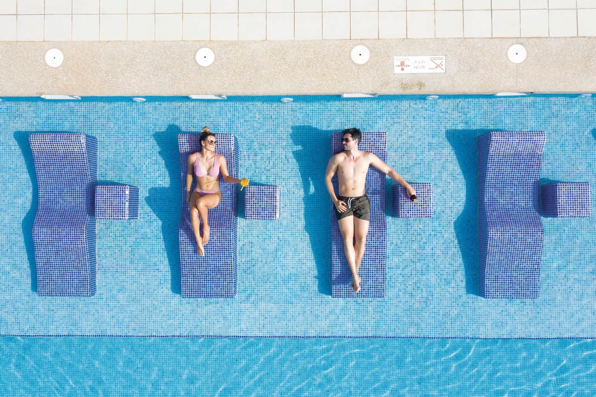 Two people enjoying drinks on a sunbed by a pool.