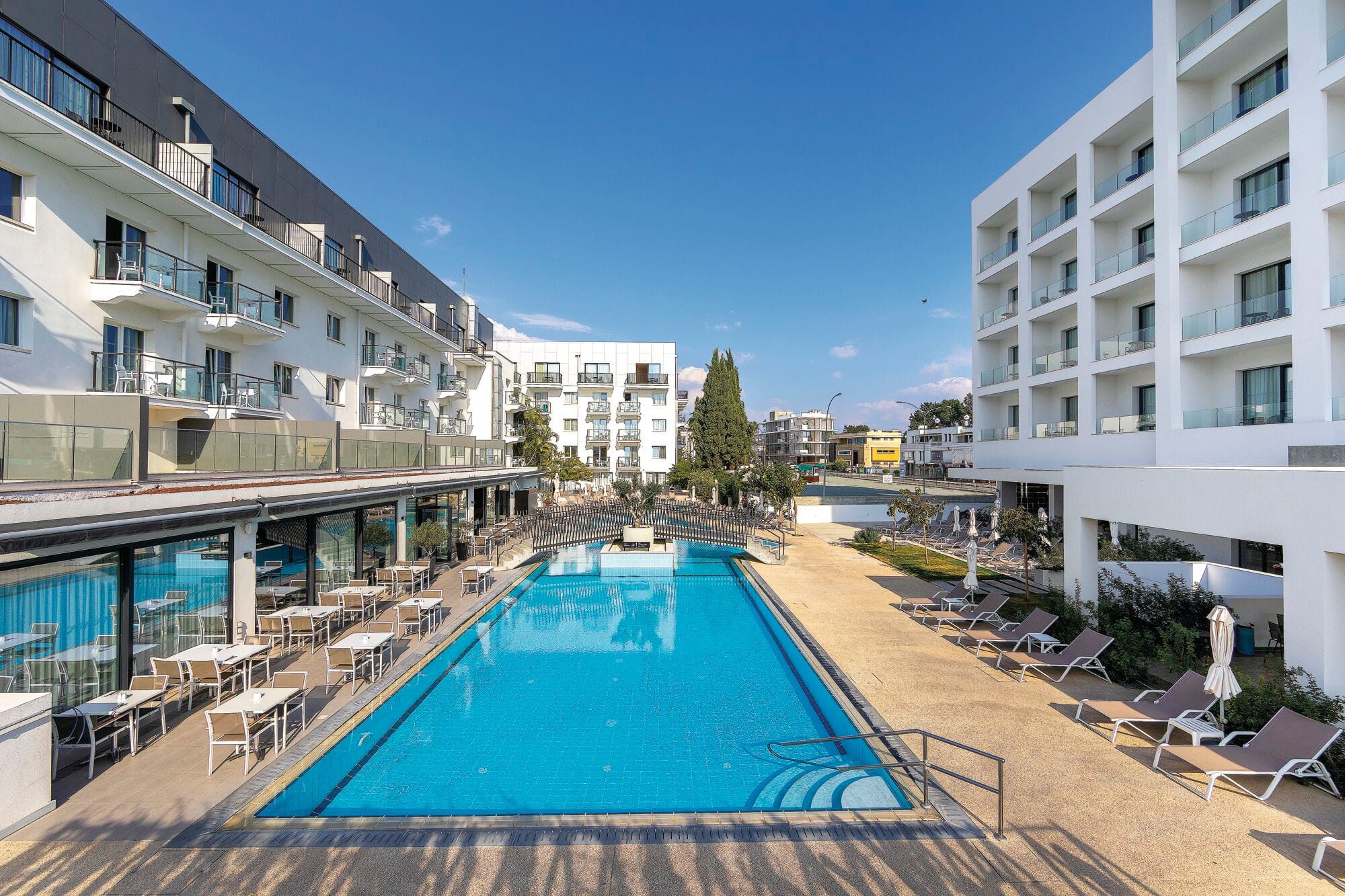 Rectangular outdoor swimming pool with bright blue water, surrounded by sun loungers and tables on both sides. Modern white multi-story hotel buildings frame the pool area, with clear blue sky in the background.