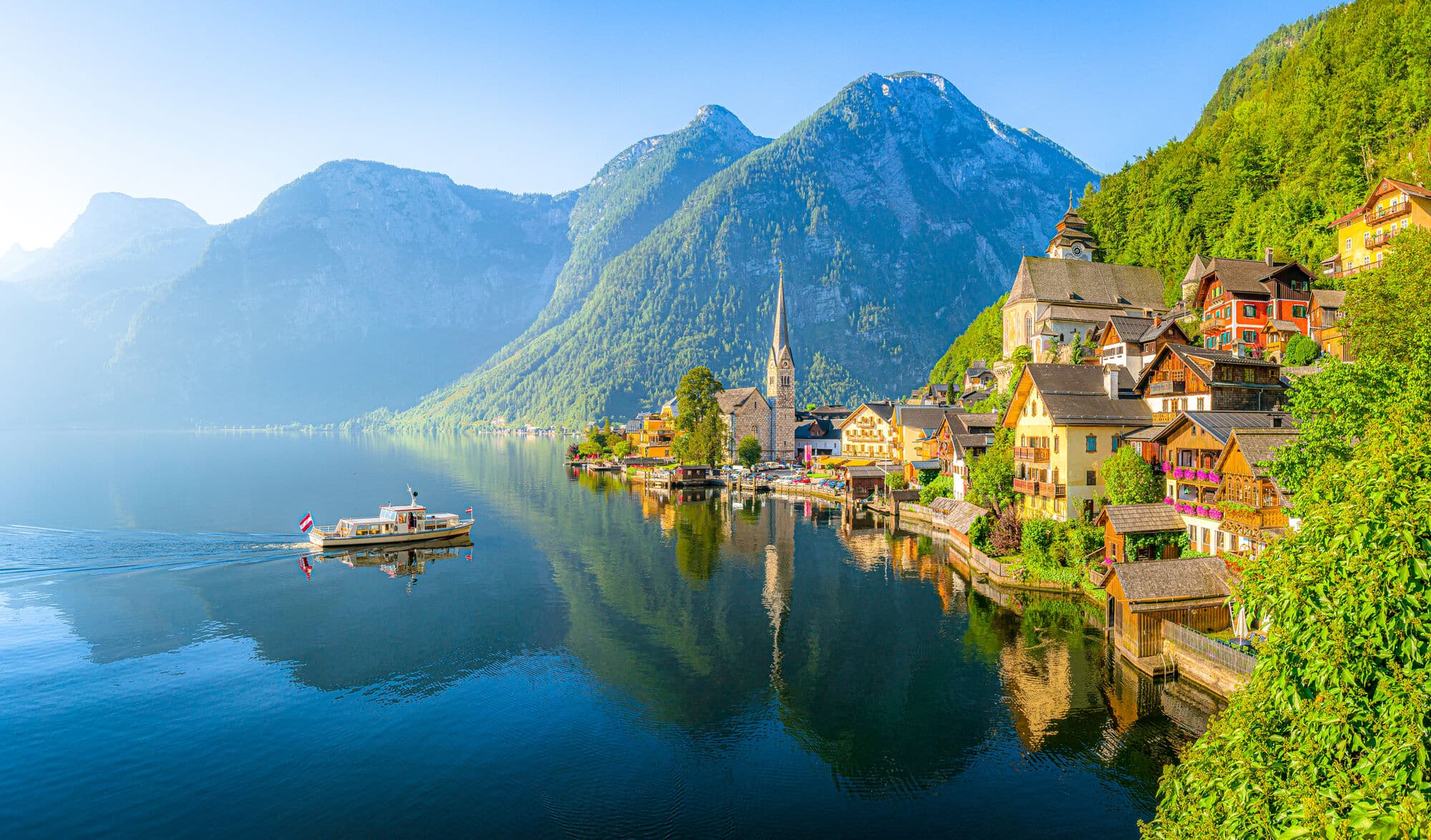 A traditional Austrian town with church spire on the edge of deep blue lake with tourist ship heading towards the town, mountains in the background