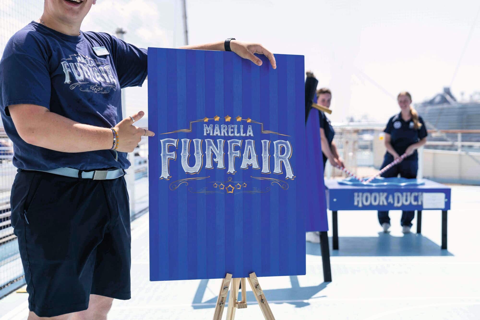 A person pointing to a Marella funfair sign, with fairground games in the background.
